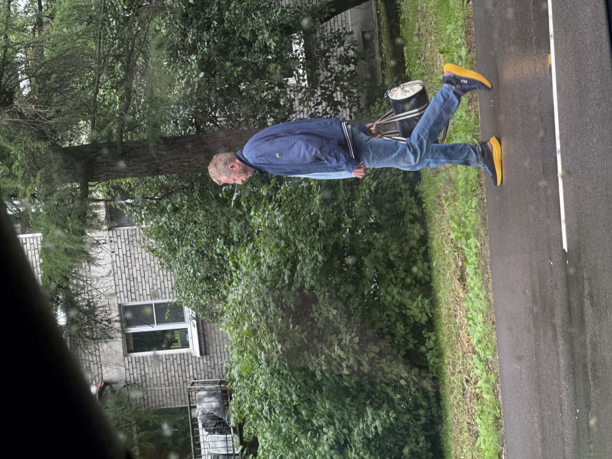 A person is walking on a wet asphalt road in Saint Petersburg, Russia. The person is wearing a blue jacket, blue jeans, and yellow and blue sneakers. They are carrying a white and black duffel bag over their shoulder. The scene is an outdoor setting with lush green trees and foliage on the left side of the road. A brick building with a window is visible behind the trees. The road appears wet, suggesting it might be raining or has recently rained. The lighting indicates it is daytime, likely overcast. There is no visible text in the image.