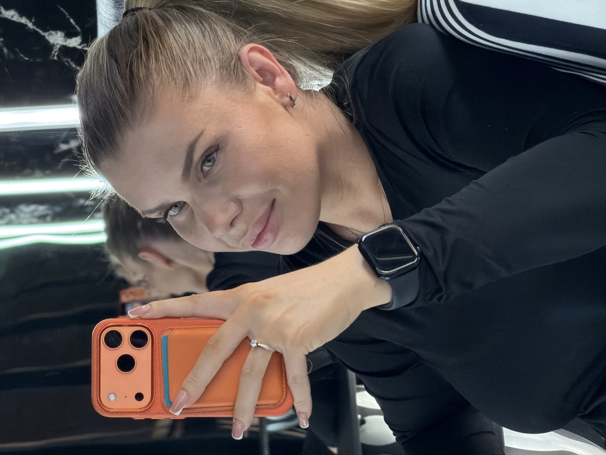 A woman is taking a selfie in a room with mirrored walls and bright vertical lights. She is wearing a black long-sleeved shirt and a black smartwatch. She is also wearing a ring on her left hand and small stud earrings. She is holding an orange phone case with a phone inside. The room appears to be a salon or dressing room. The overall lighting is dim, with the bright lights reflecting in the mirrors. There are no other people visible in the image. The location of this scene is not specified in the image itself, and therefore cannot be determined.