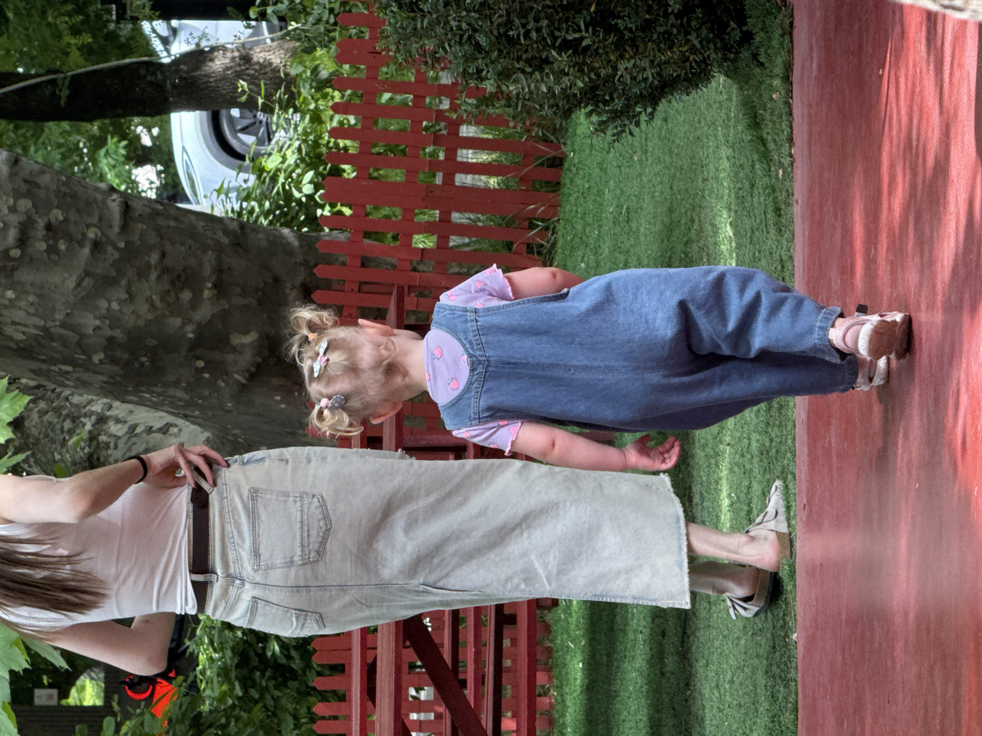 A little girl with fair skin, wearing blue denim overalls over a short-sleeved shirt with small pink strawberries on it, stands on a red surface, possibly a painted concrete or wooden patio, adjacent to a patch of green artificial grass. The girl has blonde hair styled in pigtails with small, light-colored hair accessories. She is looking down.
To her left, a taller person, likely an adult female, stands on the artificial grass. She is wearing a white t-shirt and long, light-wash denim skirt. The skirt is belted. Her hair is long and brown. She is looking at the girl.
Behind them is a low, red picket fence, separating the grass from some greenery, bushes, and a tree. A silver car can be seen between the fence and tree.
The location is Yerevan, Armenia.