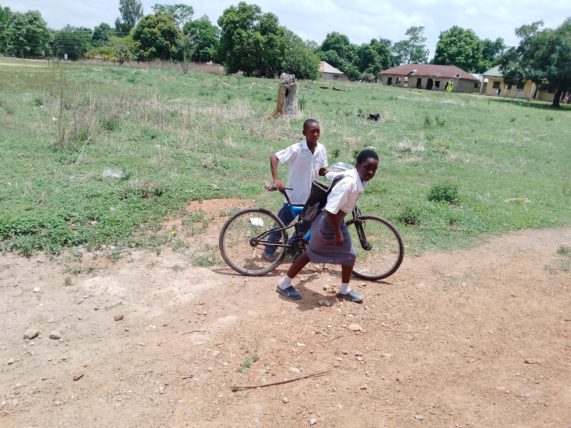 Two young people are standing on a dirt path with a bicycle. The person in the back is standing behind the seat of the bicycle and has one hand on the handlebars. They are wearing a white shirt and dark pants. The person in front is bent over and appears to be pushing the bicycle while walking. They are wearing a white shirt, a grey skirt, and white socks with dark sandals. There is a backpack on their back. The path they are on is dirt with some small rocks and sparse vegetation. Behind them is a grassy field with some trees and bushes. In the distance, there are some buildings. The location appears to be rural. The city and state are unknown.