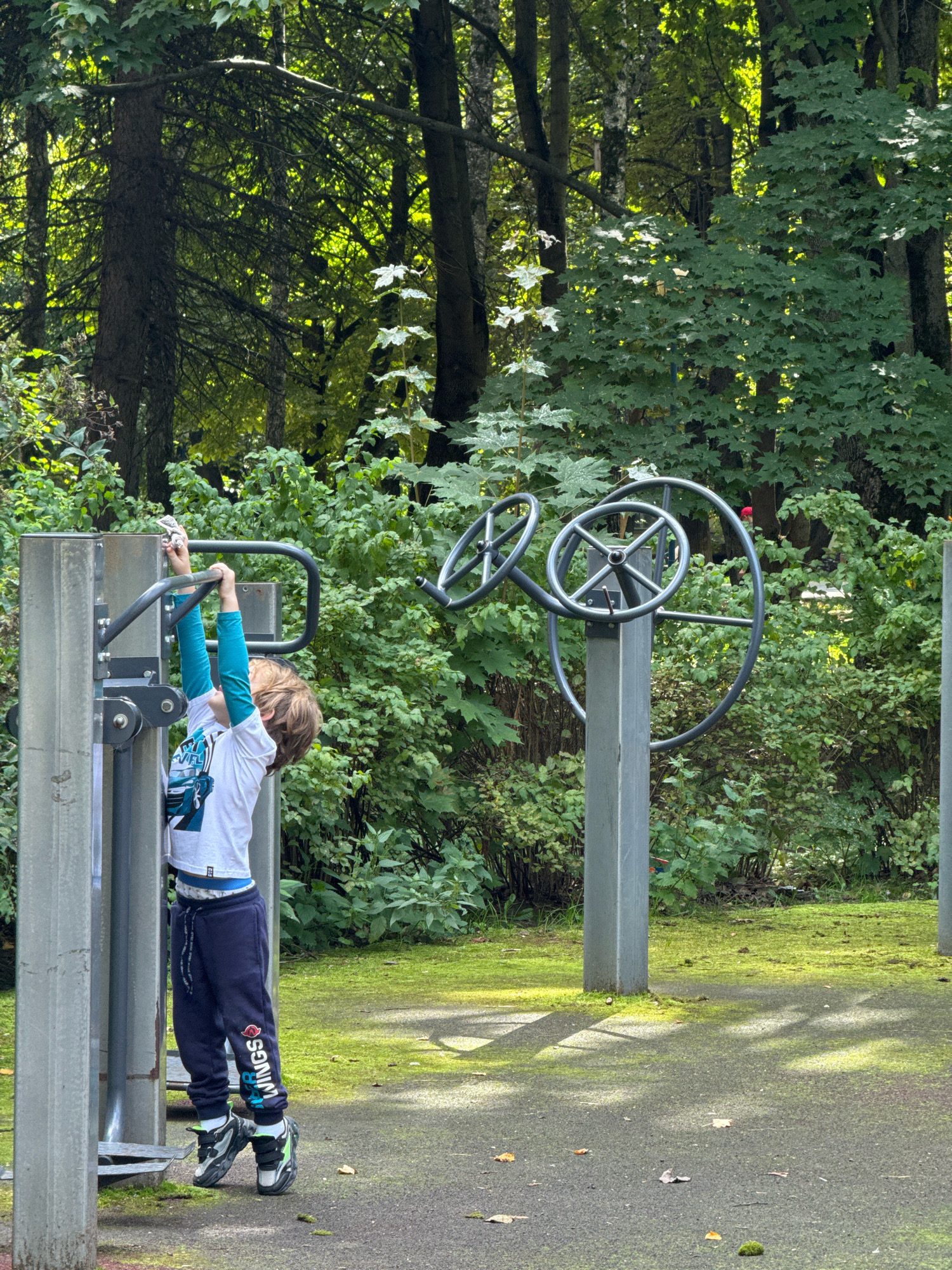 A young boy with light brown hair, wearing a white t-shirt with a blue graphic, teal long-sleeved undershirt, navy sweatpants with colorful text graphics, and black and neon green sneakers, is using an outdoor fitness apparatus in a park setting. The boy is standing on a metal platform and gripping the horizontal bars of the equipment, with his arms extended upwards. The fitness equipment consists of two gray metal posts supporting black circular hand wheels. The surrounding environment is a lush, green forest with trees and dense foliage. Sunlight filters through the trees, casting shadows on the paved ground, which is covered in patches of moss and scattered leaves. The scene appears to be in a natural, outdoor public space.