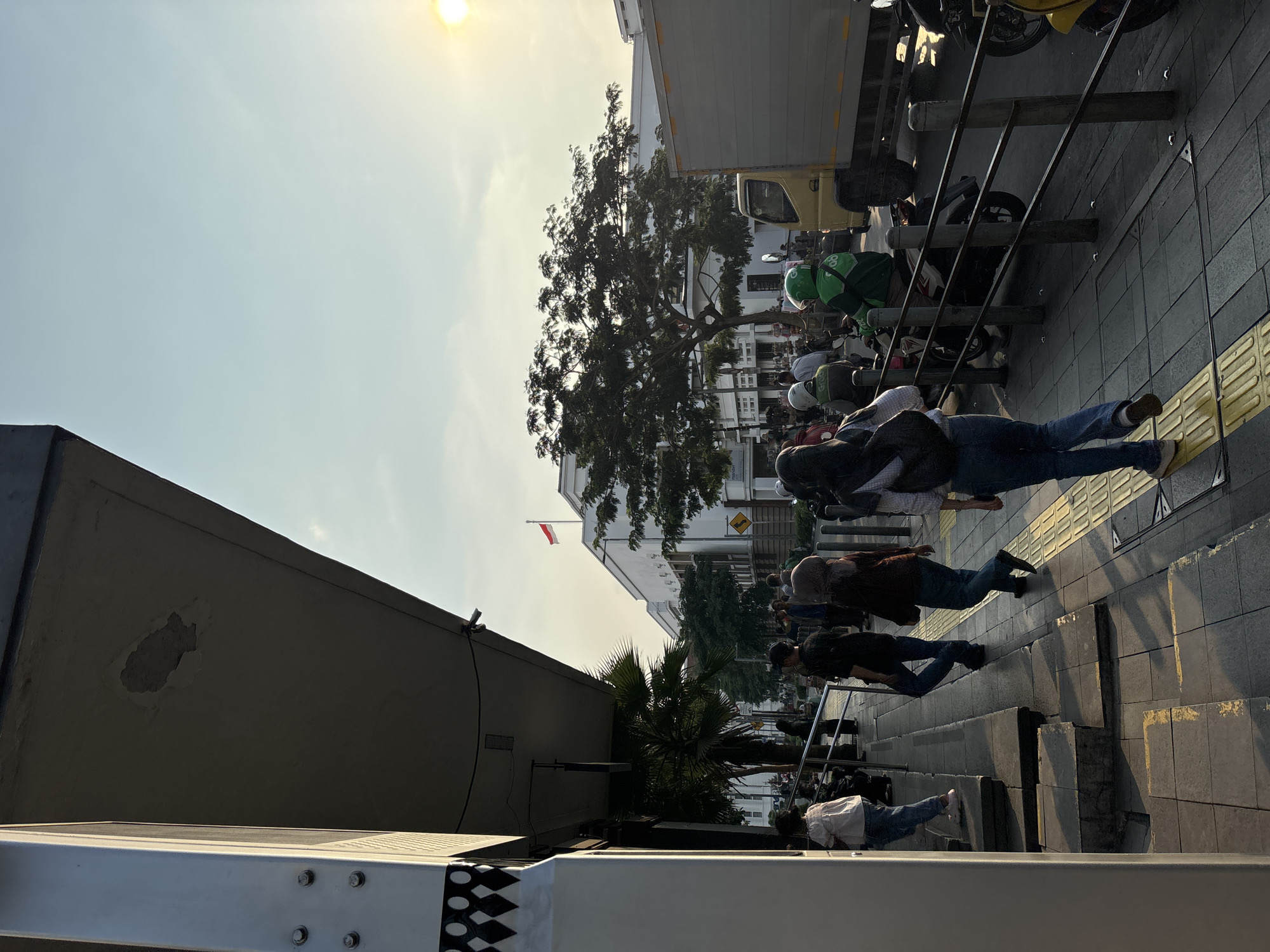 The image captures a street scene in the vicinity of Harmoni Plaza, Central Jakarta, Indonesia, during the late afternoon. The sun is low in the sky, casting long shadows across the sidewalk. Several pedestrians are walking along the sidewalk, some are waiting. On the right side of the image, a yellow truck with a grey cargo box is parked, and a line of motorcycles, some carrying passengers, are visible. The Indonesian flag is prominently displayed on a building in the background. A large tree with dense foliage stands between the building and the street. The sidewalk has a tactile paving system for the visually impaired. The overall impression is one of a bustling urban environment under warm sunlight.