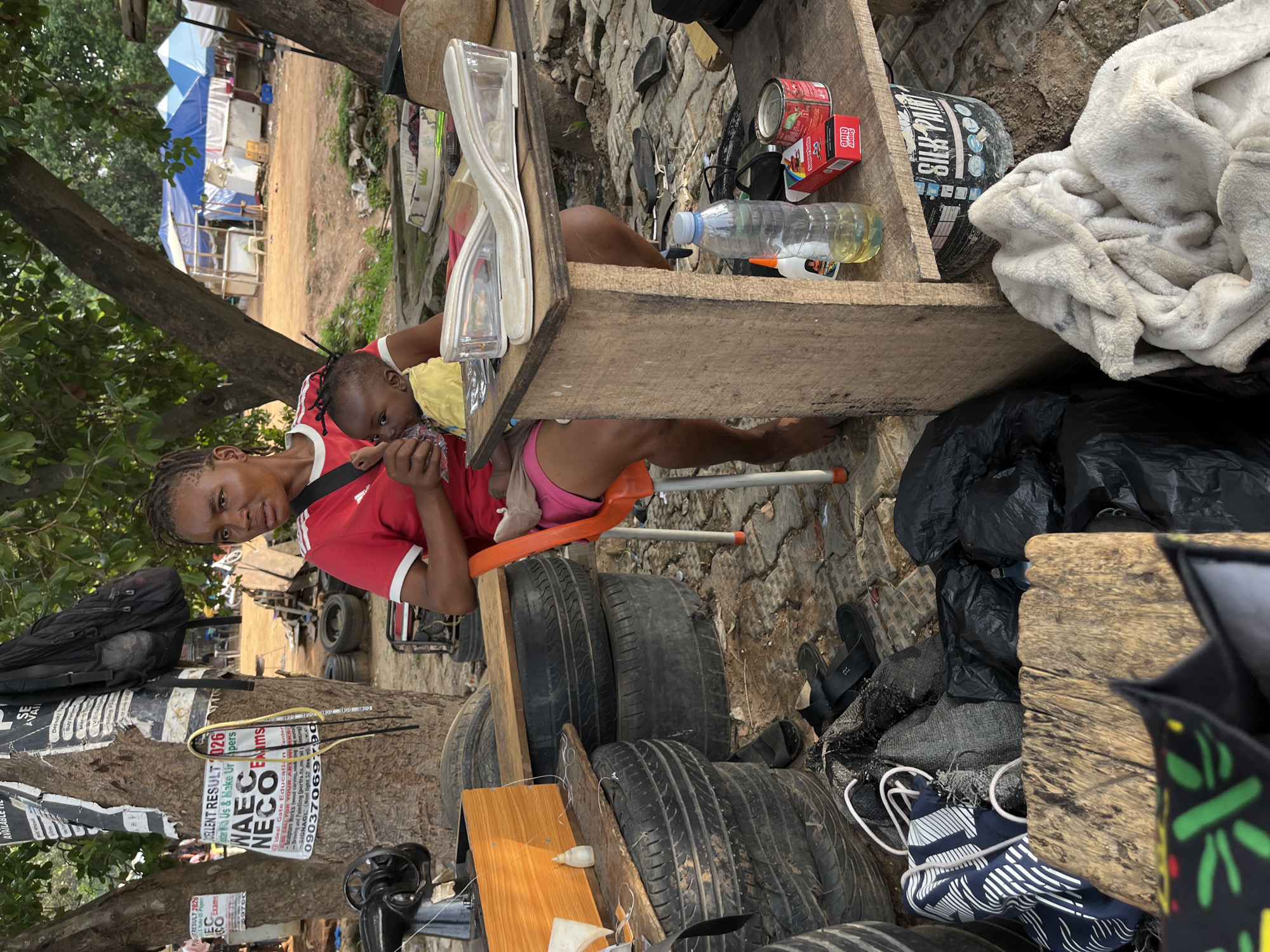 A woman sits on an orange chair, holding a baby and looking at the camera. The scene appears to be an outdoor workspace in Abuja, Nigeria, evidenced by posters on a tree advertising WAEC/NECO exams. The woman is positioned next to a makeshift table constructed from tires and wooden planks. Various objects are on the table, including shoes, glue, paint cans, and a water bottle. In the background, tents are visible, suggesting a market or similar commercial activity. Bags and other materials are scattered around the area.