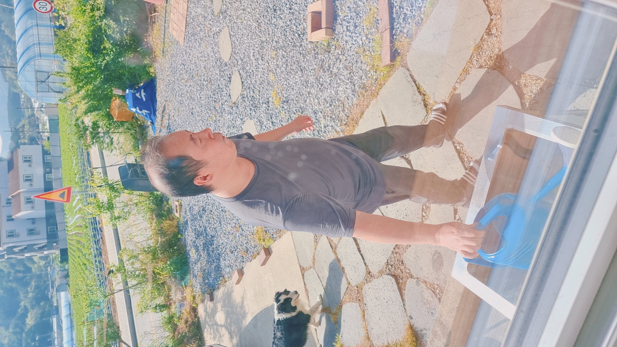 A man stands on a stone-paved area next to a house in Dunnae-myeon, South Korea. He wears a gray striped shirt and dark pants with striped footwear. He is holding a blue watering can near a window. A black and white dog is nearby. The background shows a rural landscape with houses, a street, fields, and a structure covered in clear plastic or glass. A triangular sign is visible by the road. The area is surrounded by trees and greenery.