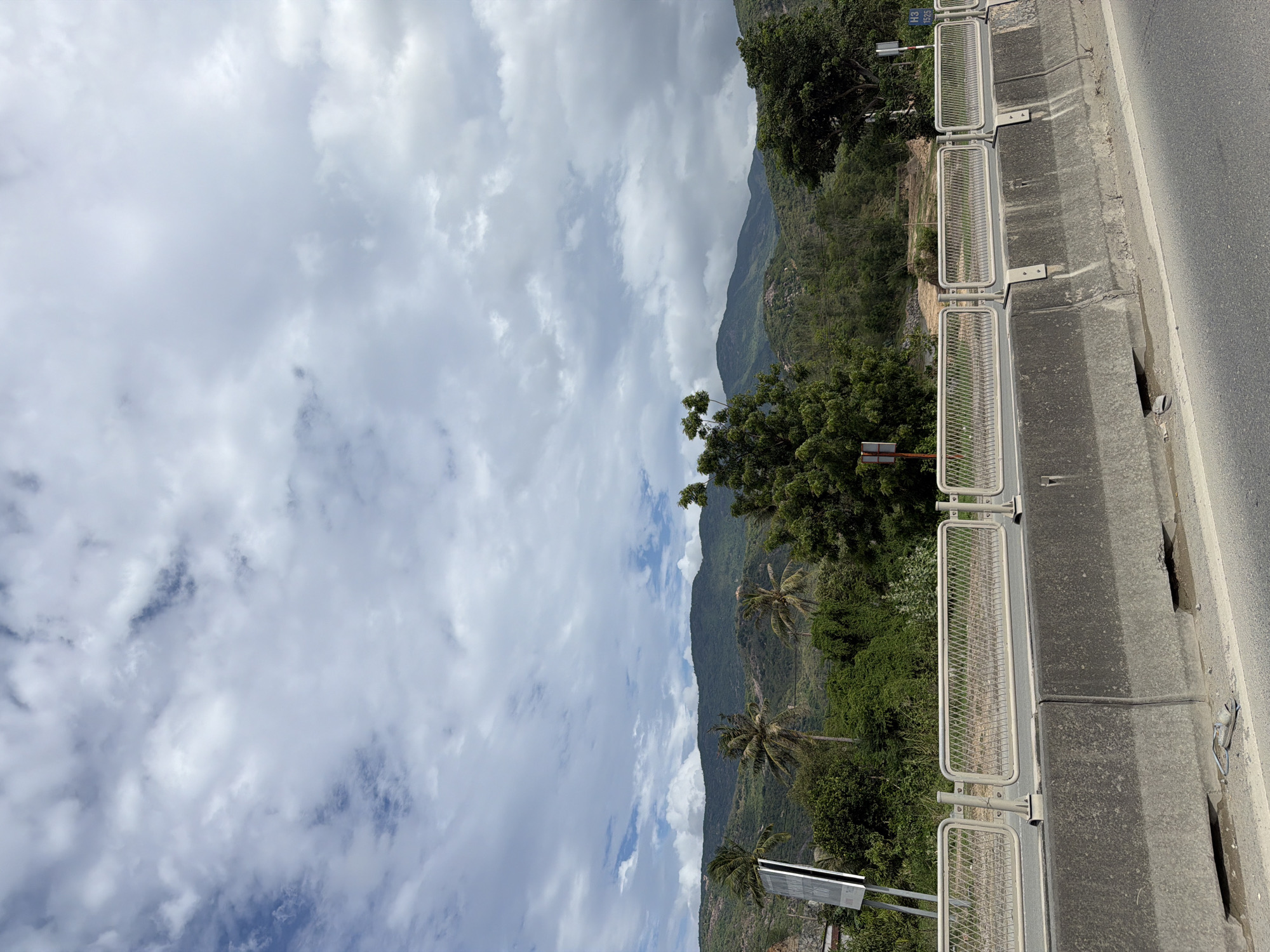 This landscape image, likely taken from a roadside perspective, captures a scenic mountainous region under a partly cloudy sky. The primary subjects include a paved road, a protective barrier system, lush green mountains, and an expansive sky.
On the right side of the frame, a section of an asphalt road is visible, bordered by a substantial concrete barrier. Resting on top of this barrier are several sections of light-colored metal guardrails, featuring a rectangular frame with a mesh infill, designed to provide safety or delineate the edge of the road.
Beyond the barrier, the terrain slopes downwards into a valley densely covered with a variety of green vegetation, including numerous trees and shrubs. Several prominent palm trees are scattered throughout this verdant landscape, particularly in the middle ground, indicating a tropical or subtropical environment. In the background, majestic green mountains rise, their undulating peaks covered in similar lush foliage, stretching across the horizon.
The sky above is filled with a mix of white and grey cumulus clouds, with occasional glimpses of blue sky peeking through, suggesting a partly cloudy day. The lighting is bright and diffused, consistent with daytime conditions.
A small white sign with blue text, partially visible near the guardrails, displays the characters "H3" and "1325", likely indicating a highway designation or a distance marker. The scene depicts a tranquil, natural environment alongside a modern road infrastructure, fitting the context of Cong Hai, Vietnam. No human activity is depicted.