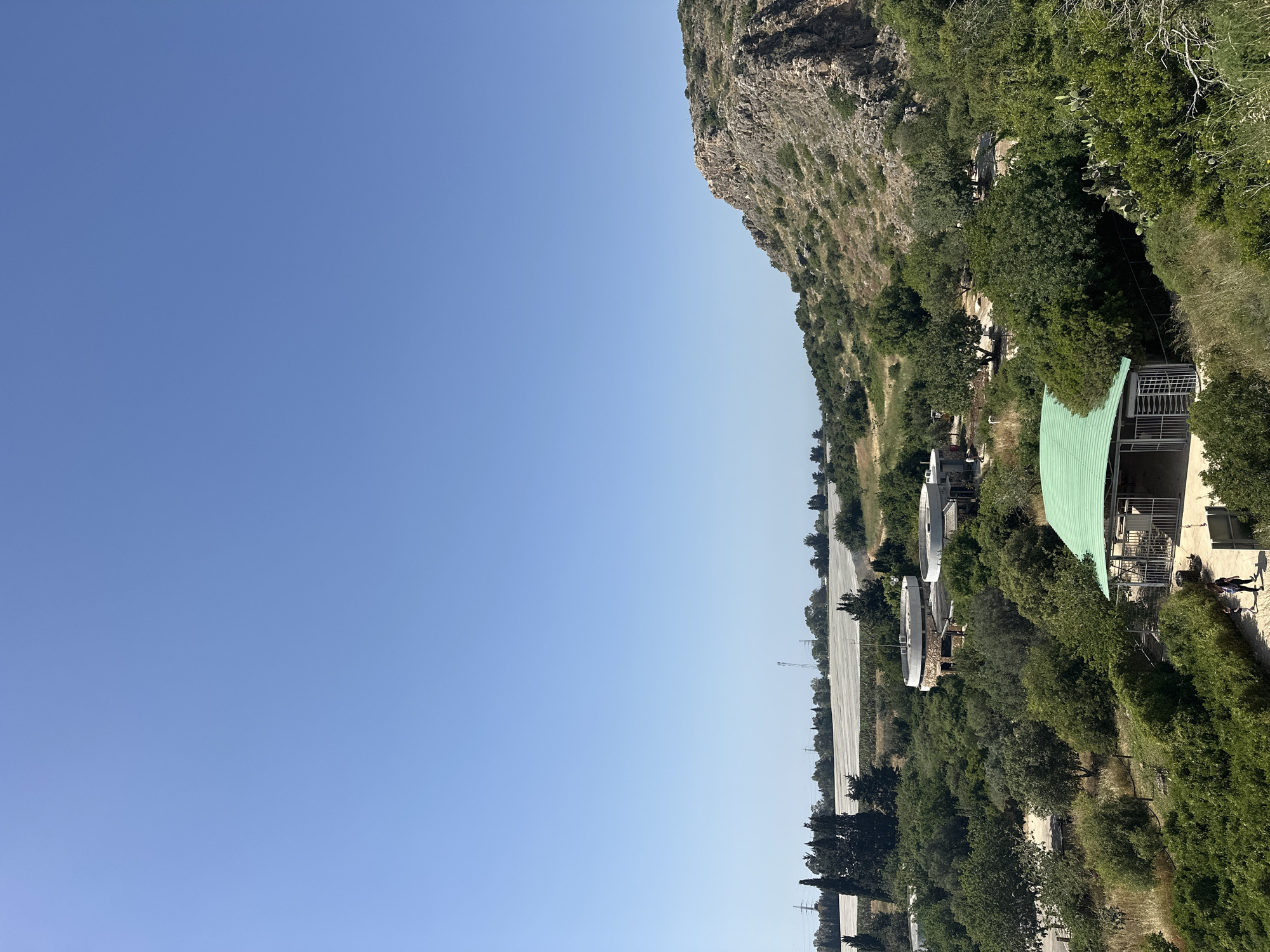 A landscape image, shot horizontally, features a clear blue sky taking up the majority of the left-hand side. A cliff face borders the sky, partially covered in green vegetation and rock. Adjacent to the cliff is a road running parallel to the cliff. There are white circular structures interspersed with trees and a green-roofed building on the right side of the road. People are walking along a path in front of the green-roofed building. The location is in the Hof HaCarmel Regional Council, Israel.