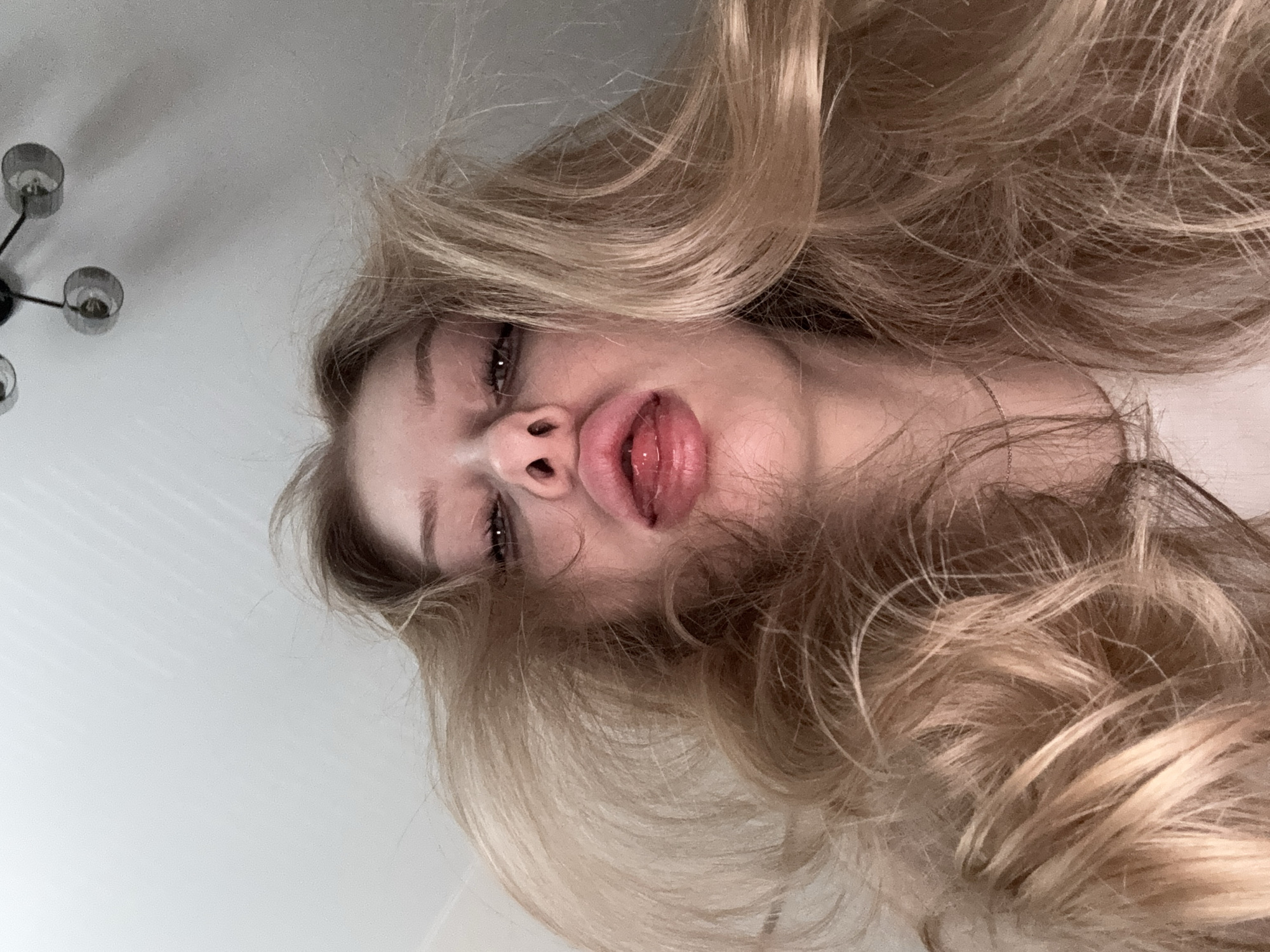 A close-up, tilted, low-angle shot of a woman with long, blonde, wavy hair. Her face is prominent, with emphasis on her lips, which are slightly parted, revealing her tongue. Her eyes are partially closed. A chandelier is visible in the upper left corner of the frame. The background appears to be a plain white wall. There is no indication of location or city.