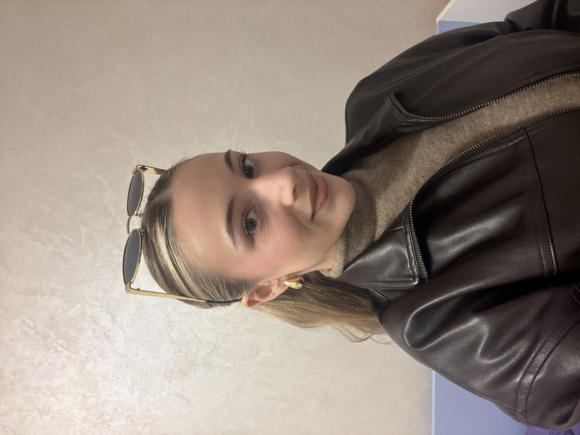 The image presents a medium close-up of a young female subject positioned against a light-colored, textured indoor wall. The subject has blonde-streaked hair pulled back into a ponytail, with gold-framed sunglasses perched on top of her head. Her facial features include light-colored eyes, defined eyebrows, a subtle blush on her cheeks, and light-toned lipstick; she exhibits a slight smile and directs her gaze forward. She is attired in a dark brown, zippered jacket worn open over a tan, high-necked sweater. Gold-toned teardrop hoop earrings are visible in both ears, and a small stud piercing is present in the upper cartilage of the subject's right ear. A segment of a light blue object is partially visible in the bottom left corner of the frame. The indoor setting provides no specific visual indicators to identify the location as the city of Plovdiv, Bulgaria.