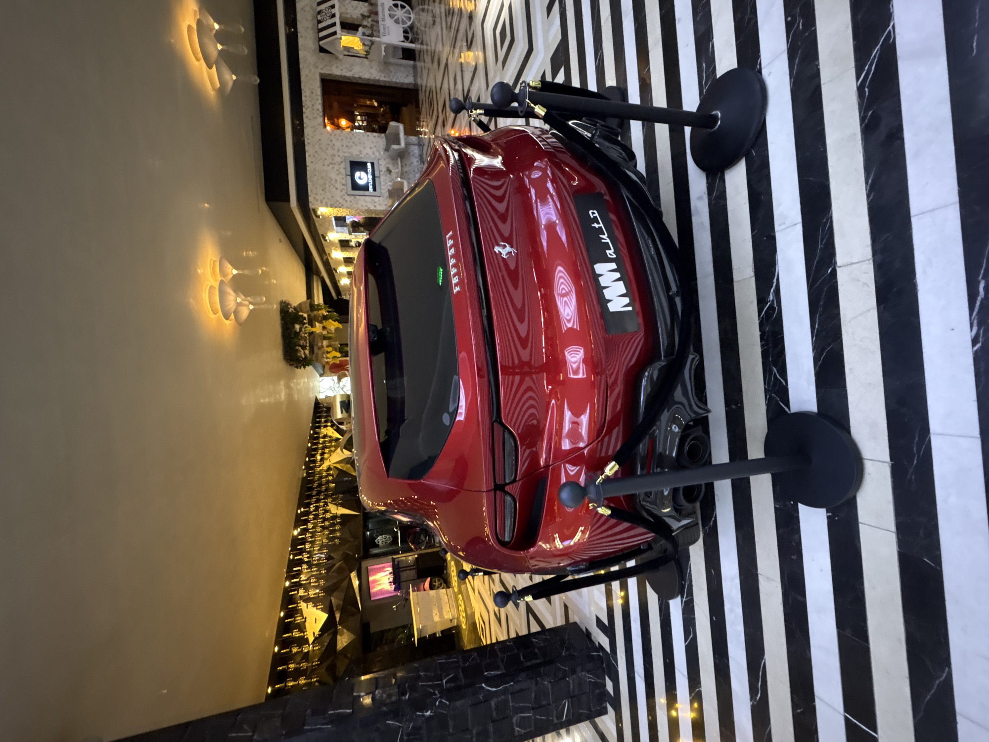 A red Ferrari car is displayed in a showroom or event space in Sofia, Bulgaria. The car is positioned on a black and white striped floor, surrounded by stanchions with velvet ropes. The environment includes mirrored walls and decorative lighting.