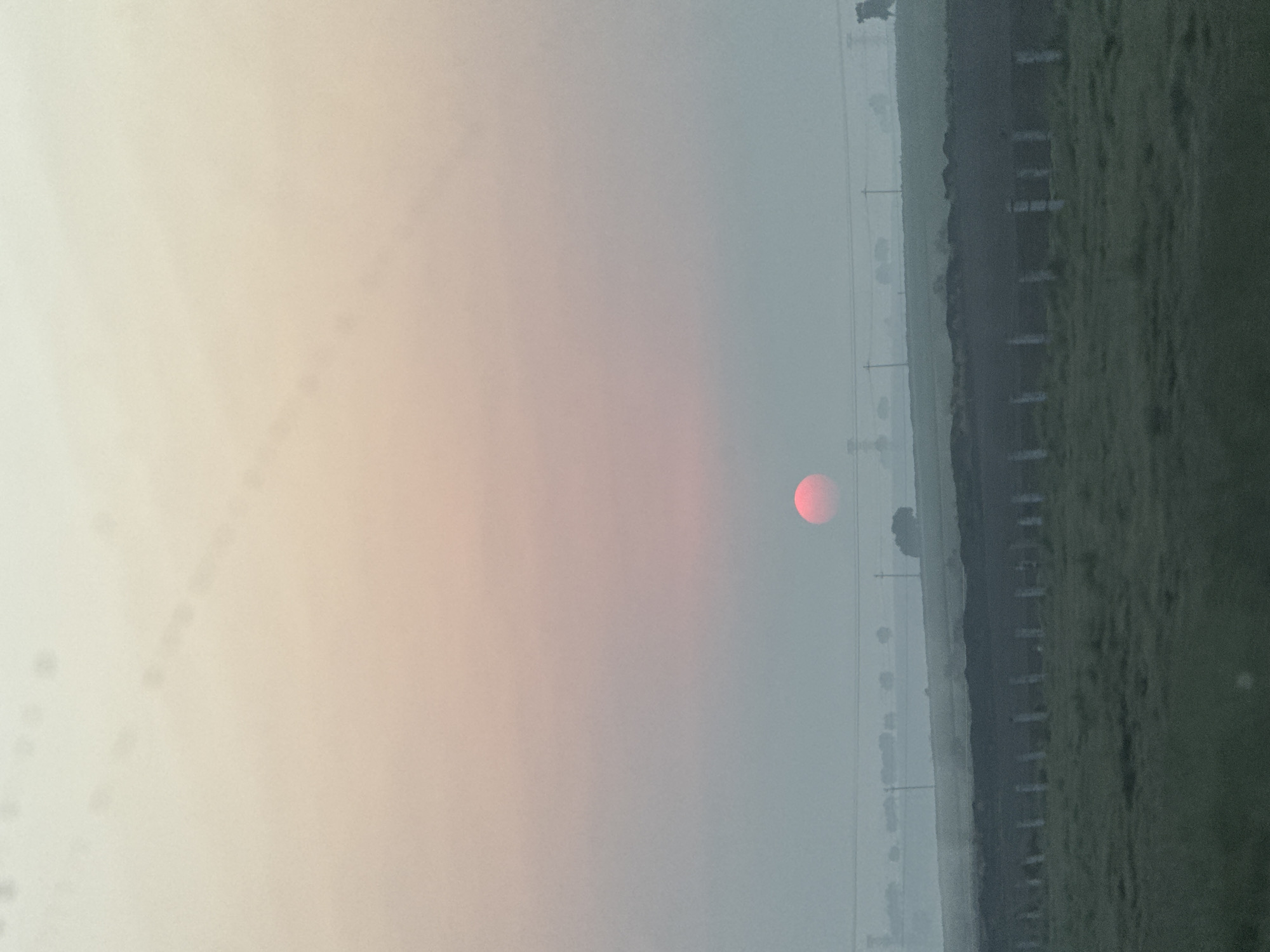 The image shows a hazy view of a rural landscape. The horizon is approximately in the middle of the frame, with the sky occupying the upper portion. A red sun is visible on the horizon, obscured by the haze. The lower portion of the image shows a field with a fence running along its edge. Electrical lines are visible in the distance. Based on visual cues, the picture likely shows the area around Máximo Serdán, Mexico.