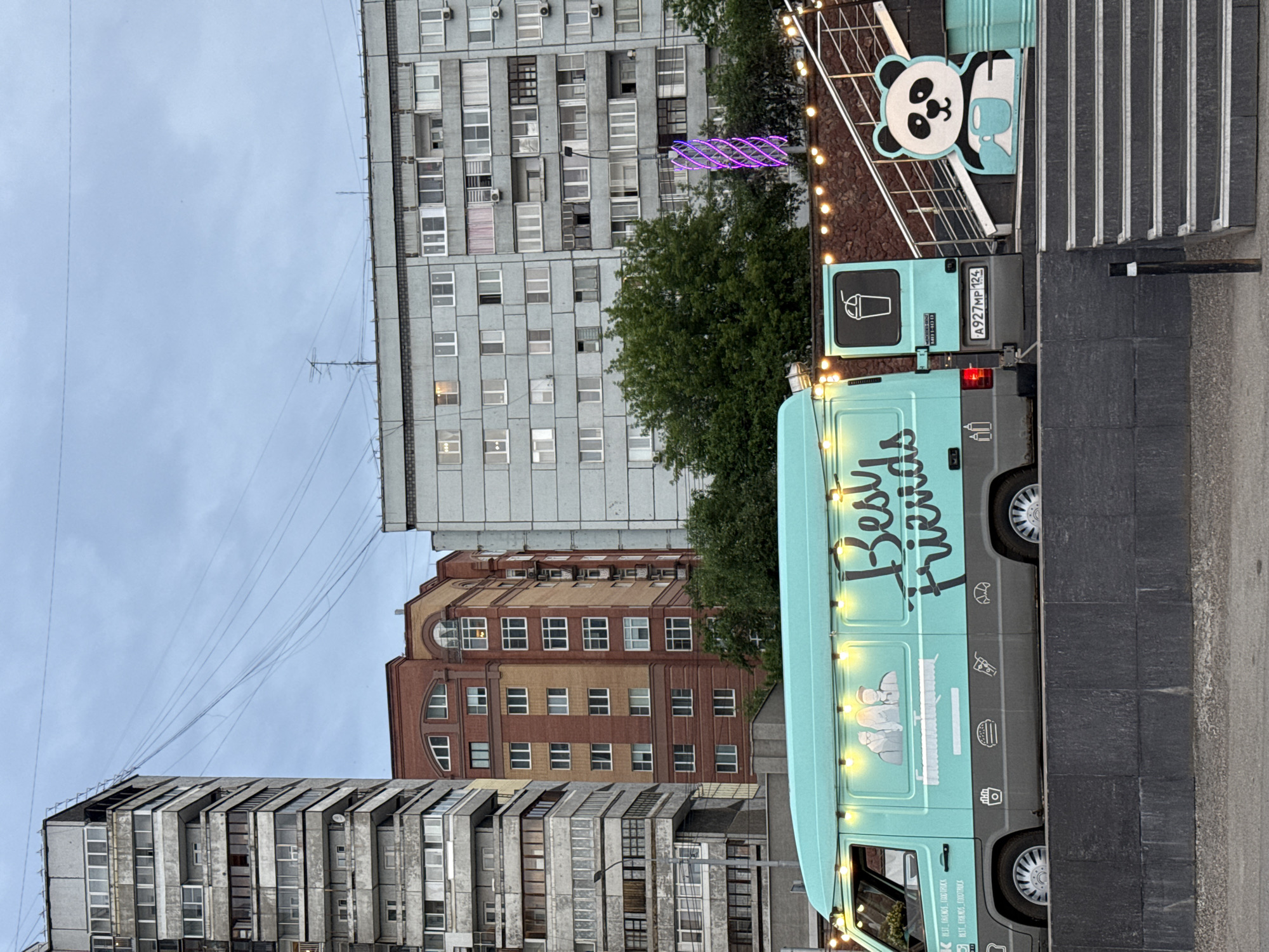 An outdoor scene features a turquoise food truck named "Best Friends" parked on a raised, dark gray platform. The truck is adorned with string lights and graphics depicting cartoon characters and food items. To the right of the truck, a staircase leads upwards, bordered by a large cutout of a panda holding a mug. In the background, several multi-story apartment buildings of varying architectural styles dominate the cityscape under an overcast sky. The environment appears to be an urban setting in Krasnoyarsk, Russia, as indicated by the license plate on the food truck, which reads "A927MP 124", consistent with Russian license plates. The overall scene suggests a commercial or recreational area within the city.