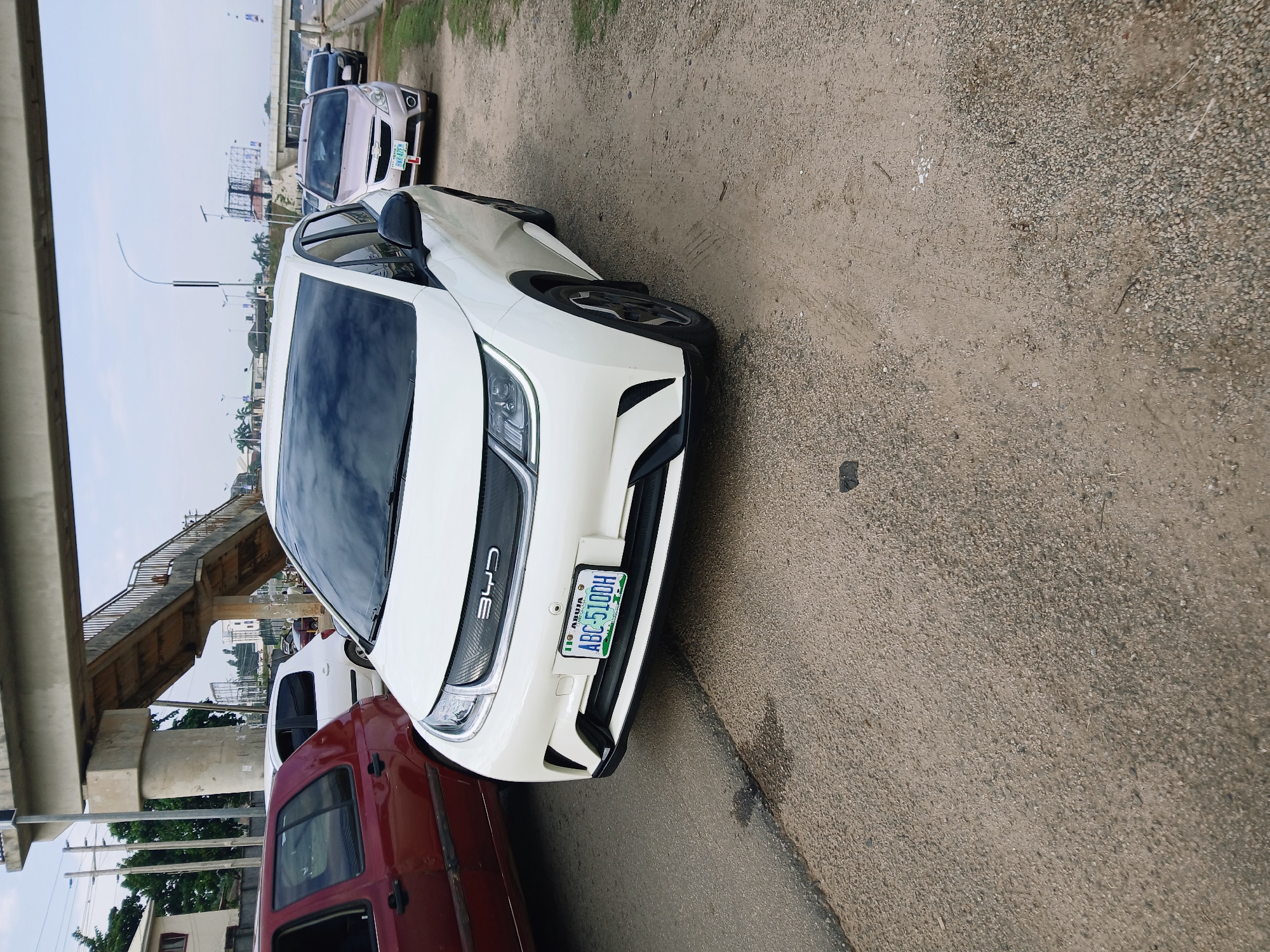 The image shows a white BYD car parked on the side of the road with a red van next to it. In the background, there's a pedestrian bridge, other vehicles, and buildings. The license plate on the BYD car reads "ABC-510DH" and indicates it is from Abuja. The location is presumably somewhere in Abuja, Nigeria, given the license plate information.
