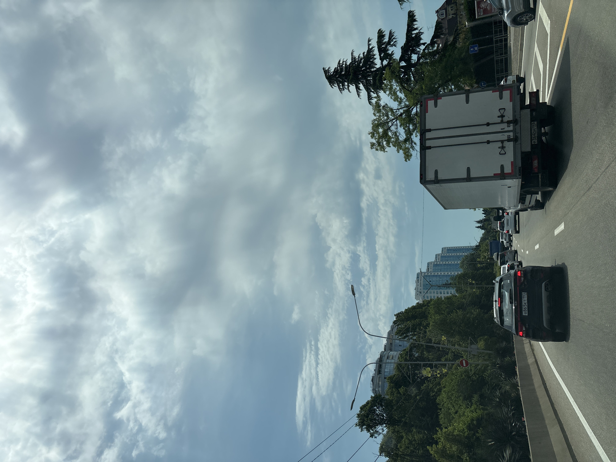 The image depicts a street scene in Sochi, Russia, likely during daylight hours with overcast skies. The perspective is from a vehicle traveling on a multi-lane road. In the foreground, the rear of a black Kia SUV is visible, its license plate reading "A865KT 193". To the right of the SUV, a white box truck is in motion, its rear doors closed. Further ahead, other vehicles, including a blue truck, are on the road.
The left side of the road is lined with lush green trees and vegetation, with several modern, multi-story buildings visible in the background. These buildings appear to be apartments or hotels, characterized by their sleek, curved architectural designs and numerous windows. The right side of the road features more trees, including a tall evergreen tree, and glimpses of buildings and signs, one of which appears to be a traffic sign with a blue square and a white symbol. Power lines are also visible in the upper left portion of the image. The road itself is paved, with clear lane markings. The overall impression is of a busy urban environment with a significant presence of greenery.