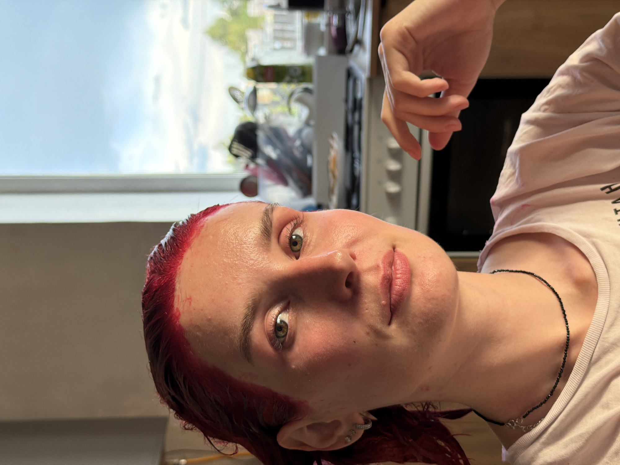 A person with red hair dye applied is facing the camera. The person has fair skin and hazel eyes. They are wearing a light pink t-shirt and a black beaded necklace. The background includes a window with a partial view of foliage and sky. A white oven and kitchen appliances are also visible behind them. The location or its relevance to the city of Chaikovsky, Russia is not ascertainable from the image.