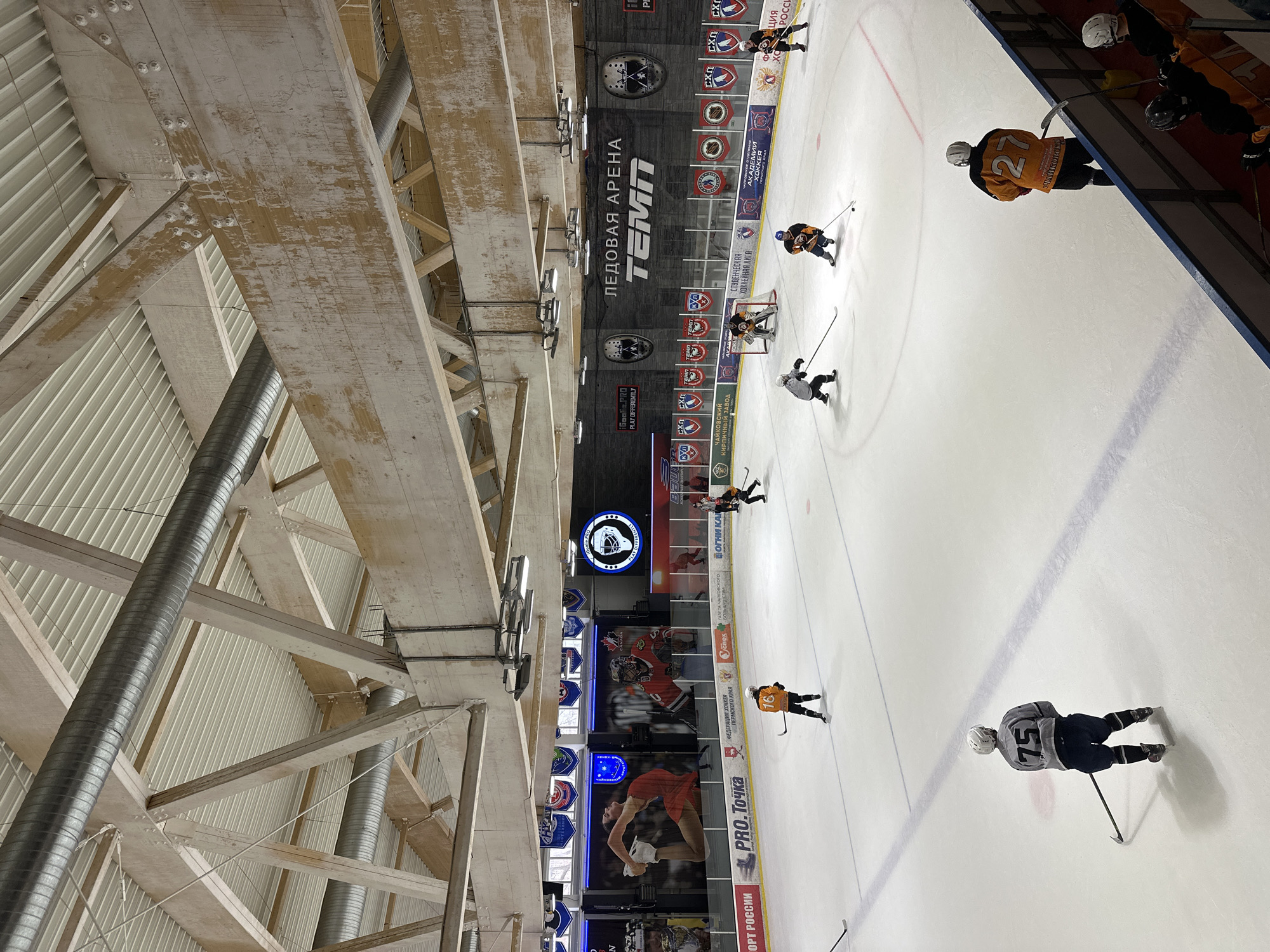The image captures a hockey game in progress at the "Ledovaya Arena Temp" (Ice Arena Temp) in Chaikovsky, Russia. The arena features a prominent banner with the arena's name, along with logos and advertisements for various organizations. Several hockey players, identified by numbers "75" and "27" are on the ice. The arena's ceiling is exposed, revealing a network of beams, pipes, and ventilation systems. Advertisements for "Pro.Tochka", "Federation of Hockey Perm region", "Gazeta Tchaikovsky" are visible on the boards.