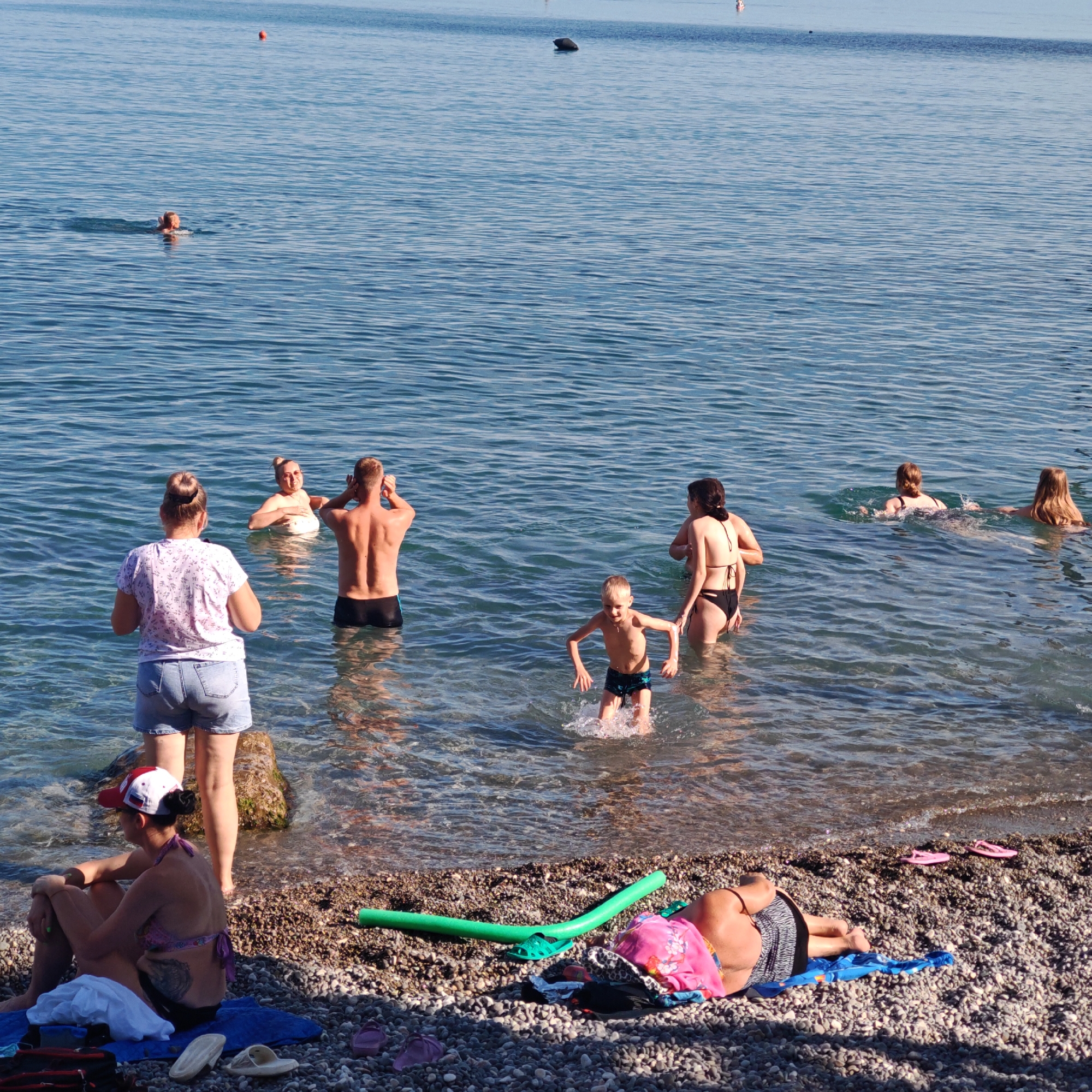 In the foreground, a woman sits on a pebble beach, wearing a patterned bikini top and a red and white cap. She is looking to her left. Next to her, a person stands in the shallow water. A green pool noodle rests on the beach.
In the water, several people are enjoying the day. A man stands with his hands on his head, facing away from the camera. A woman in a white top stands in the water behind him, looking towards the man. A young boy is splashing in the water in front of them. Further out in the water, a woman in a black bikini is swimming. Two other women are also swimming in the distance. There are buoys in the background, indicating a swimming area.
The scene is a beach on a sunny day, likely late morning or early afternoon given the bright sunlight. The water is calm and clear, and the overall atmosphere appears relaxed and recreational. The location is by the sea, consistent with a beach resort. No text is visible in the image.