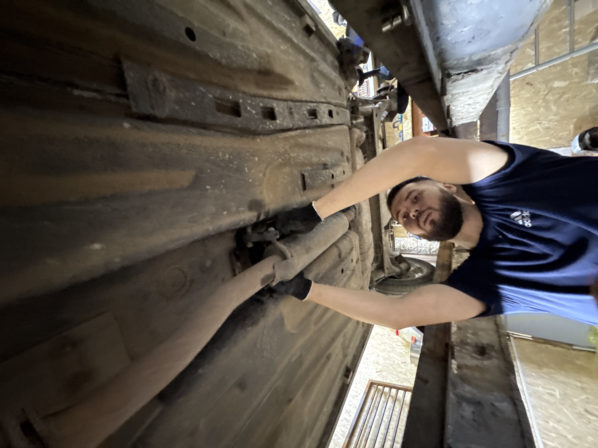 A bearded man, wearing a blue Adidas t-shirt and black fingerless gloves, is captured from a low angle, looking upwards with focused attention. His arms are raised, and his gloved hands are actively engaged with the underside of a vehicle, specifically manipulating a section of the exhaust pipe, which appears dirty and slightly corroded. The car's undercarriage is visibly soiled with dirt and grime, showing areas of surface rust on its metallic components and structural frame rails. The vehicle is significantly elevated, positioned either over a service pit or on a mechanical lift, with heavy, dark-colored support beams prominent in the foreground and mid-ground, exhibiting some wear and peeling paint. The background reveals the interior of a workshop or garage, characterized by walls clad in light-colored plywood or OSB panels. To the left, a metal grate structure is visible. To the man's right, beneath the elevated car, various tools, including a socket set, are arranged on a workbench, with a partial view of a ladder further in the background. This scene depicts automotive maintenance being performed in a garage in Perm, Russia.
