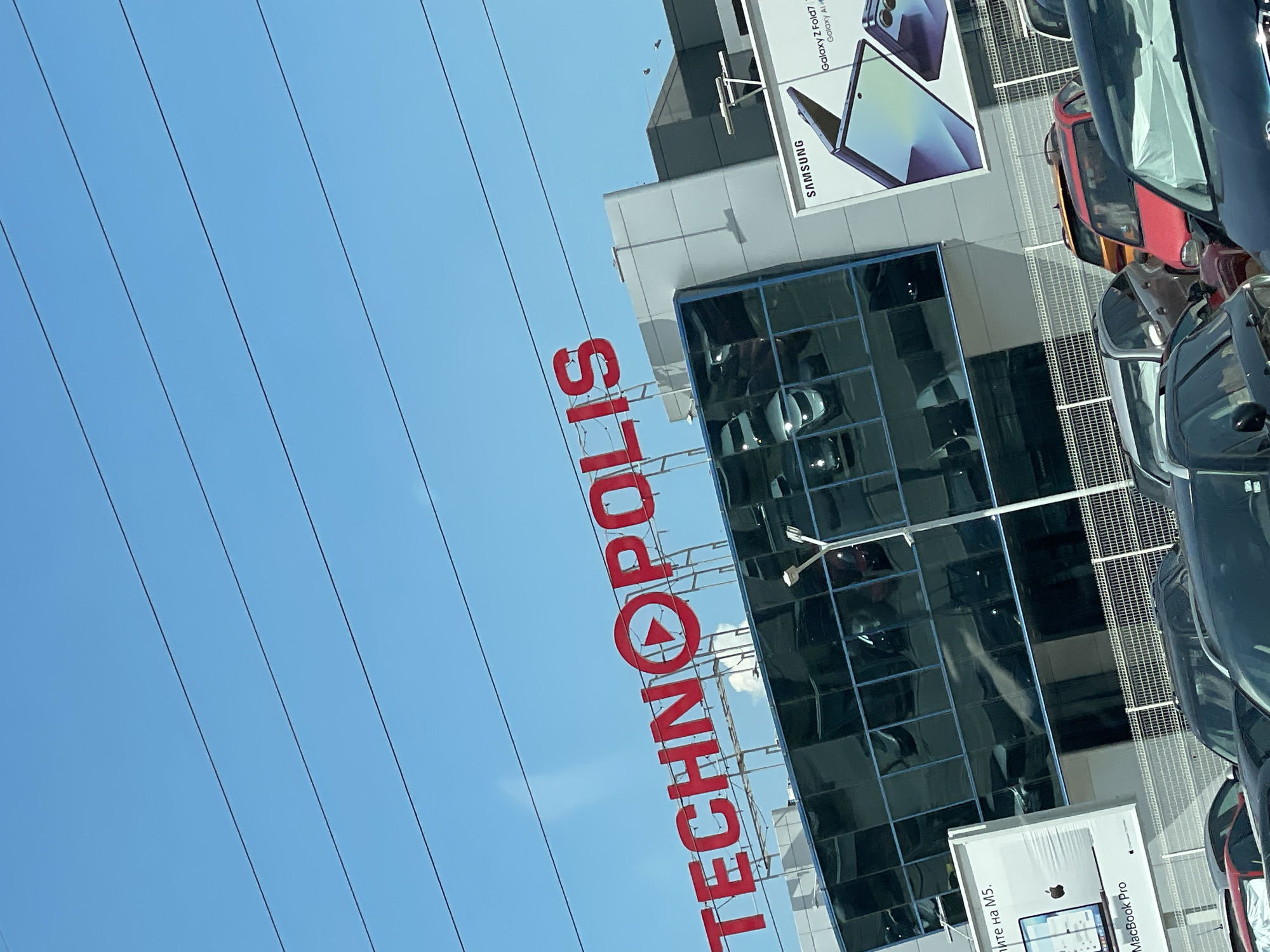 The image shows the exterior of a Technopolis store in Sofia, Bulgaria, on a clear, sunny day. The store features a large red Technopolis logo with a play button icon. The building has a modern design with large, dark tinted glass windows reflecting the sky and surrounding environment. A billboard advertising Samsung products, including the Galaxy Z Fold, is visible on the right side of the building. In front of the building, there is a parking lot filled with various cars. The sky is bright blue with visible power lines crossing the frame diagonally. The image appears to be taken from a moving vehicle, likely a car, judging by the perspective and slight blur.