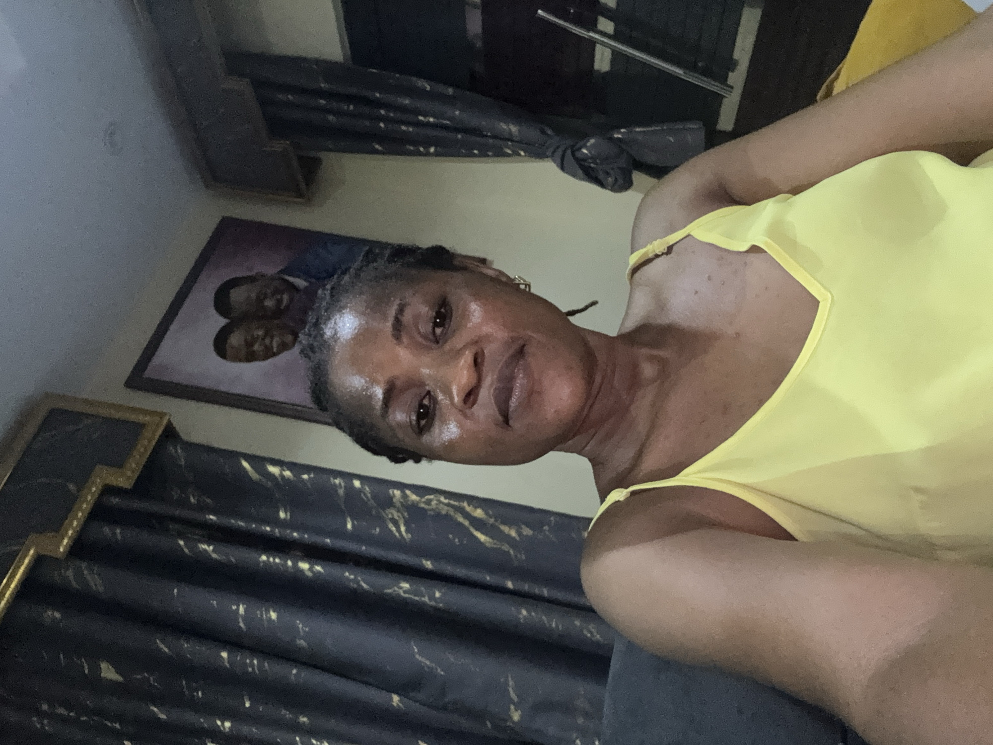 This media file shows a close-up, selfie-style shot of a Black woman. She is wearing a bright yellow tank top, and her hair is styled in braids. She appears to be indoors, with dark curtains featuring a gold, abstract pattern visible in the background. A framed portrait of two men in suits is also visible behind her. The lighting suggests it could be daytime, but the indoor setting makes it difficult to determine the time of day precisely. The woman's expression is relaxed and direct, as if she is posing for the photo. There are no visible signs or text in the image. The overall impression is a personal snapshot taken in a home environment in Ajara, Nigeria.
