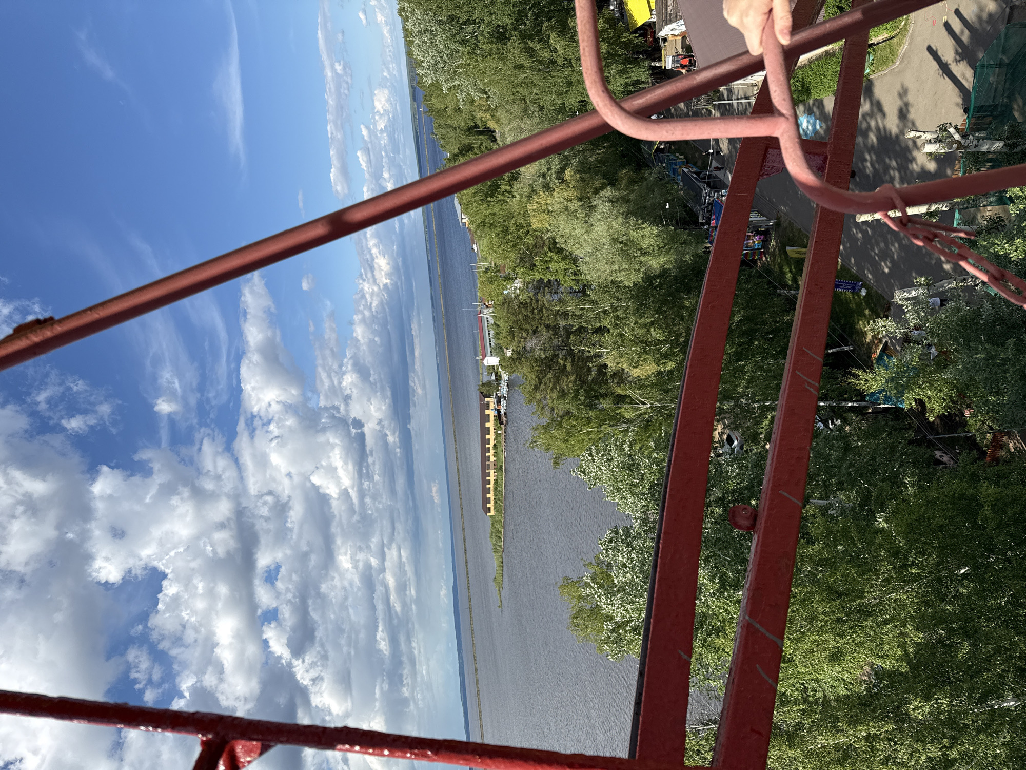 The image shows a view from a red metal structure overlooking a body of water and a park. The water appears to be a wide river or a lake, with land visible on the opposite shore. The park has trees, a road, and some buildings. The red metal structure in the foreground is likely part of an observation tower. The sky is blue with white clouds. The location is likely the observation tower in Chaikovsky, Russia. A hand is visible on the upper right.