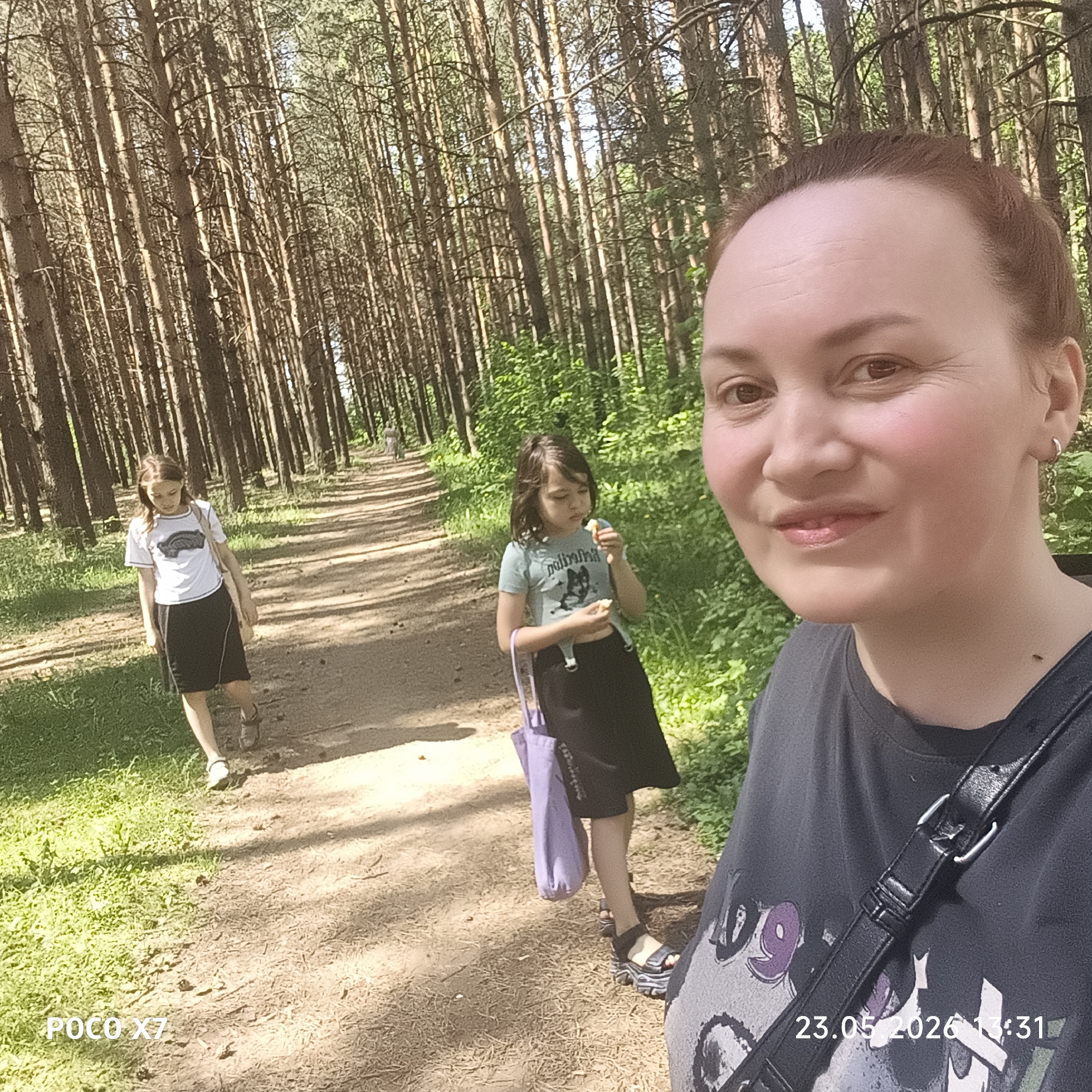 On May 23, 2026, at 13:31, a person is taking a selfie on a path in a forest in Chaikovsky, Russia. Two girls, possibly daughters, are walking on the path ahead. The person taking the selfie is wearing a dark t-shirt and a black crossbody bag. The girl closest to the camera is eating something. The other girl has a light-colored bag over her shoulder. The forest consists of tall, slender trees. The ground is covered with dirt and fallen pine needles, with some patches of grass. The sky is visible through the treetops. A woman is visible further away on the path. The date and time are stamped on the bottom right corner of the image.