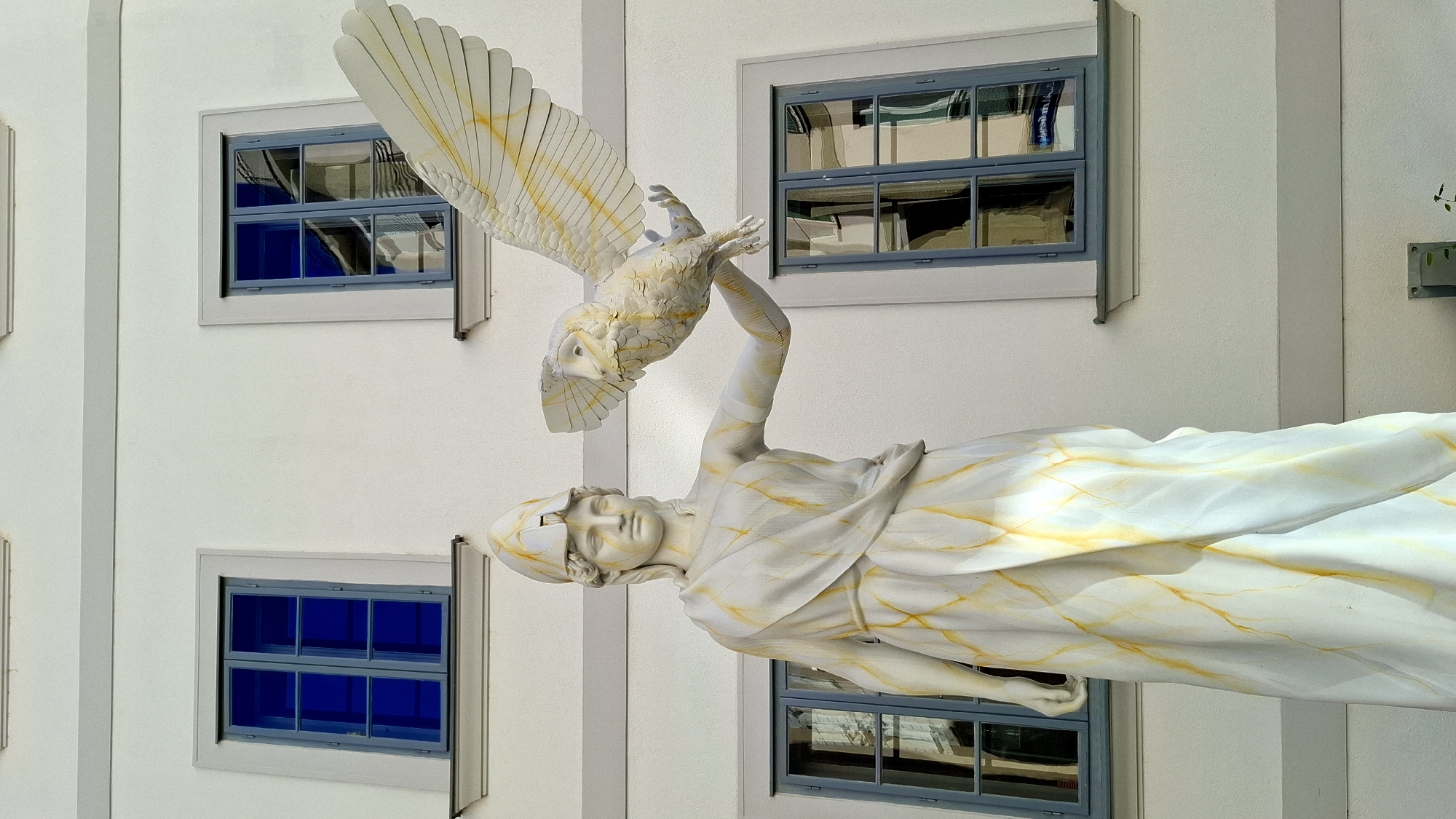 This is a full-body, eye-level outdoor shot of a white marble statue of Athena, the Greek goddess of wisdom, holding a white owl with outstretched wings. The statue is positioned in front of a white building with four rectangular windows. The statue is adorned with yellow streaks resembling lightning or veins, and the owl has similar yellow markings on its wings. The windows are framed in grey and have blue glass panes, reflecting the surroundings. The building appears to be in a European city, likely Vienna, Austria, given the architectural style and the typical appearance of such statues in public spaces there. The statue's left arm is extended, and the owl is perched on its outstretched hand. The statue is wearing a classical Greek helmet and a draped robe. The background is a plain white wall with some grey horizontal trim. The lighting suggests it is daytime, with sunlight casting subtle shadows on the statue.