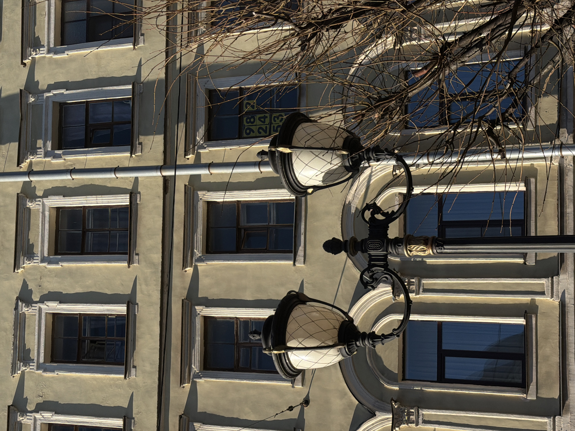 An eye-level, medium shot presents the exterior of a building in Saint Petersburg, Russia, partially obscured by a streetlamp and bare tree branches. The building's facade is light olive green, adorned with a pattern of rectangular windows framed with white molding. The windows reflect the sky, giving them a blue hue. Some windows display adhesive notes with numbers.
A black, ornate streetlamp with two white globe-shaped lights is positioned in front of the building, slightly off-center. Bare tree branches intermingle with the streetlamp and upper portion of the building. A gray pipe runs vertically alongside the building's facade. The overall tone of the image is neutral, with natural lighting casting shadows on the building's surface.