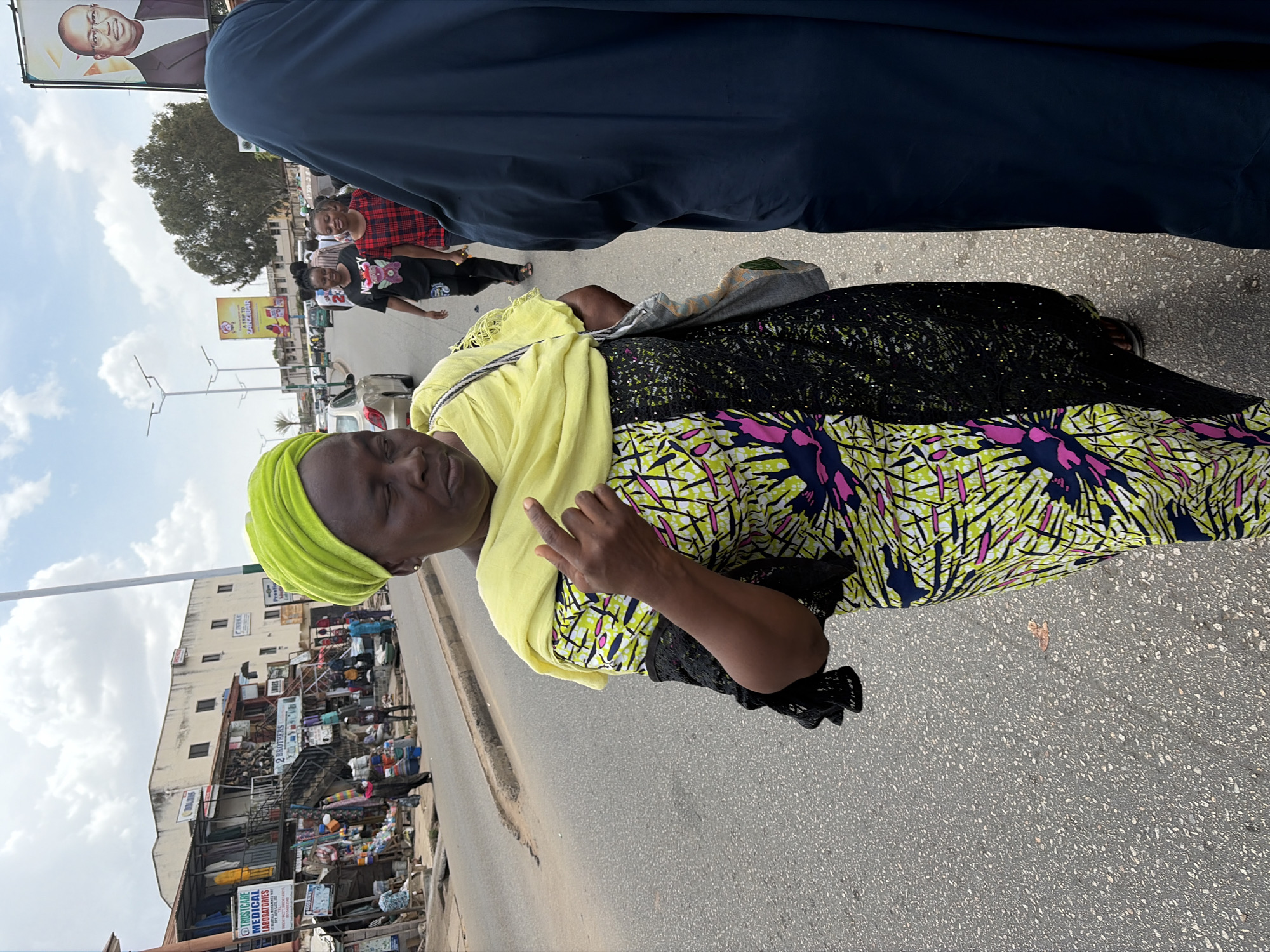 The image presents a vibrant street scene in Jos, Nigeria, during the daytime under a partly cloudy sky. In the foreground, a woman with dark skin is the primary subject, positioned facing left. She wears a bright lime green headscarf, a pale yellow shawl draped over her shoulders, and a long, eye-catching dress. The top part of her dress is black with subtle shimmering details, while the lower section features a bold pattern of lime green, pink, and dark blue abstract shapes. Her left hand is raised, with the index finger pointing upwards. Her facial expression appears calm and contemplative.
To her right, partially obscuring the view, is the back of another person dressed in a dark blue garment. Further back in the midground, several other people are visible walking and standing on the asphalt road. One woman with a red and black plaid shirt smiles towards the camera, while another person wears a black t-shirt with a white graphic.
The street itself is a busy urban environment, flanked by multi-story buildings that appear to house shops or businesses, with various goods displayed outside. High above, on the upper left, a large billboard features the portrait of a smiling man in a suit, likely a political figure or a public personality. Other visible text includes "Jumia Pay ZANZIBAR" on a yellow billboard in the midground, and a sign for "TRUSTCARE MEDICAL LABORATORIES" nearer the bottom left. Trees and utility poles are also scattered throughout the background, completing the impression of a lively and active cityscape.