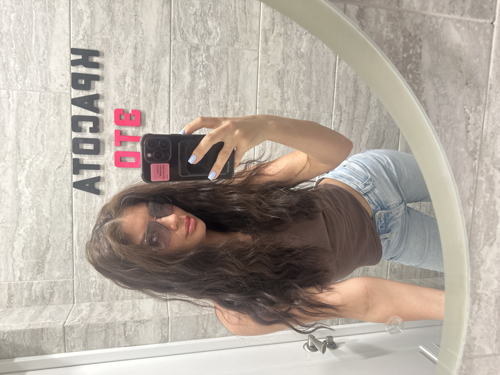 The image is a mirror selfie of a young woman taken in an indoor setting, likely a bathroom or vanity area, characterized by tiled walls and a sink. The woman is positioned to the left of the frame, capturing her reflection in an oval or circular mirror that occupies the right side of the image.
The main subject is a woman with long, wavy, dark brown hair. She is wearing a dark brown, ribbed crop top and light blue, distressed jeans. She has on large, rectangular sunglasses with a metallic frame. Her lips appear to have a subtle pink or nude color. Her right hand, with light blue painted fingernails, holds a black smartphone to take the photo. A pink sticker or label with indistinct text is visible on the back of her phone case.
The background consists of light-colored, greyish, marble-patterned wall tiles. Below the mirror, a white sink basin and a metallic faucet fixture are visible.
A notable detail on the tiled wall, reflected in the mirror, is a set of decorative block letters. The black letters, when read correctly (reversed from the mirror's reflection), spell "КРАСОТА" (KRASOTA), which means "beauty" in Russian. Adjacent to these are pink letters that, when reversed, spell "ЭТО" (ETO), meaning "this" or "is" in Russian. Together, the text reads "ЭТО КРАСОТА" ("This is beauty"). The lighting suggests an indoor setting, possibly with natural light or bright artificial lighting. The woman's expression is neutral, consistent with posing for a selfie.