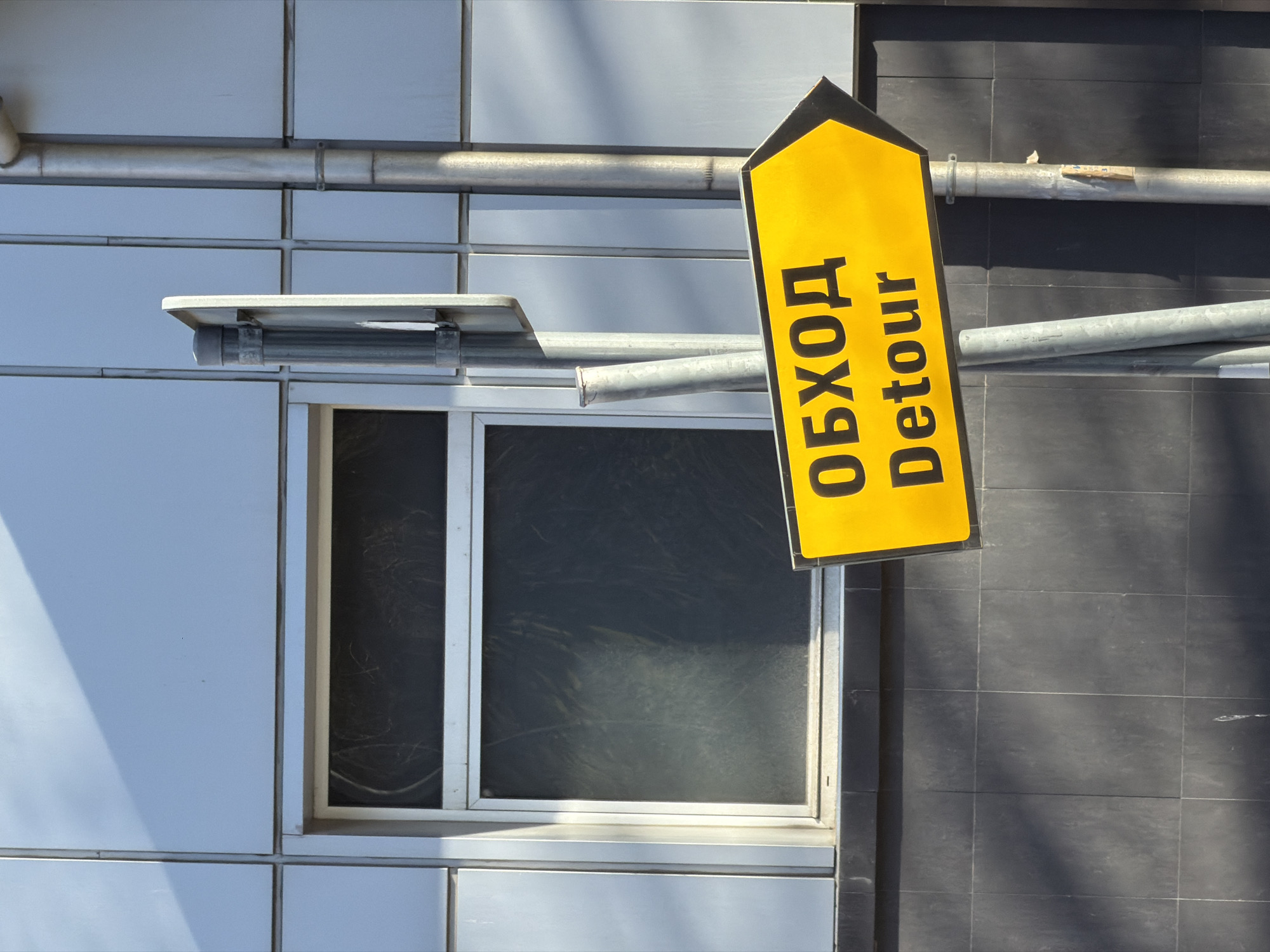 The image shows a yellow "Detour" sign, with the Cyrillic word "ОБХОД" above it, mounted on a metal pipe affixed to the side of a building. The building has a modern facade composed of light gray panels and dark gray tile sections. A window is visible to the left of the sign, with a white frame. The pipes appear to be utility conduits. The photo was taken in Plovdiv, Bulgaria.