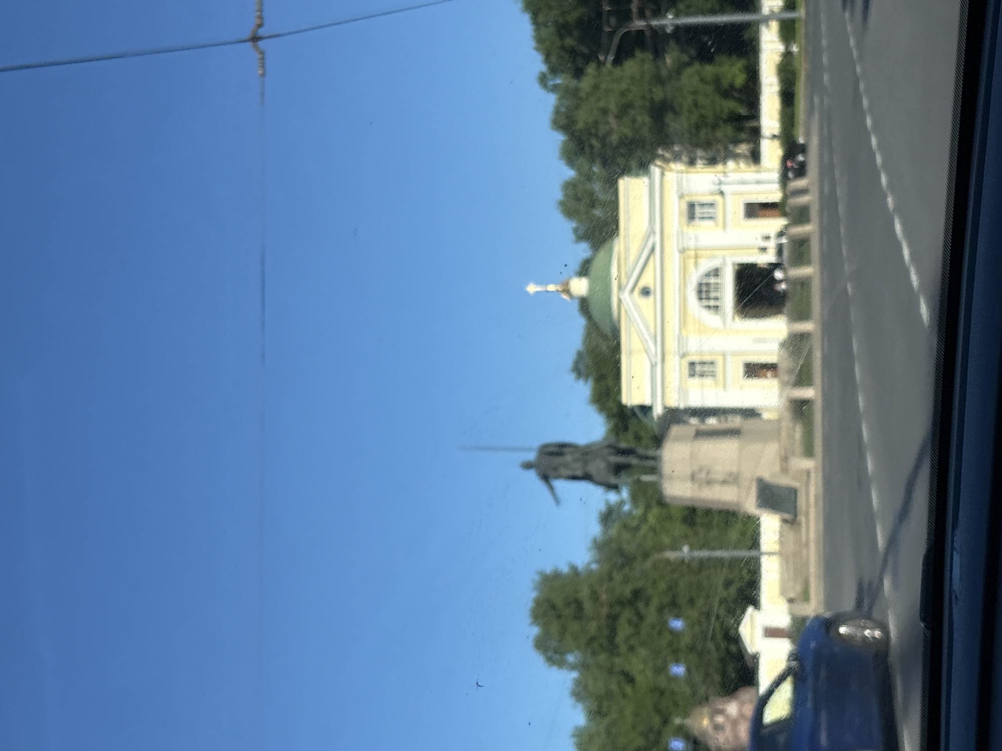 This image depicts an outdoor scene in Saint Petersburg, Russia, specifically at Alexander Nevsky Square (Ploshchad Aleksandra Nevskogo), featuring the Alexander Nevsky Monument and parts of the Alexander Nevsky Lavra. The shot is taken from the interior of a vehicle, evidenced by the dark dashboard visible at the bottom and minor obstructions on what appears to be a windshield.
The upper half of the image is dominated by a clear, bright blue sky, exhibiting no clouds. In the midground, an equestrian statue of a man on horseback, identified as the Alexander Nevsky Monument, stands on a multi-tiered, light-colored stone pedestal. The statue, appearing dark (likely bronze), shows the figure holding an upright spear or sword, extending his right arm forward.
To the right and slightly behind the statue is a light yellow, neoclassical-style building, part of the Alexander Nevsky Lavra complex, likely the Gate Church of the Annunciation. This building features a prominent arched entrance in its center, flanked by rectangular windows on two levels, and is topped by a green dome crowned with a golden Orthodox cross. A low, light-colored wall or fence runs horizontally in the mid-distance behind the building.
Dense green trees form a backdrop behind the statue and the building. A two-lane asphalt road with dashed white lane markings occupies the foreground. On the far left of the road, the front section of a dark blue car, including its wheel, is partially visible. Further left, behind the blue car, a smaller, darker domed structure with a cross is partially visible through the trees. Overhead wires are visible crossing the upper right portion of the sky. Minor dark specks are scattered across the image, consistent with dust or smudges on a glass surface. The overall focus of the image is slightly soft.