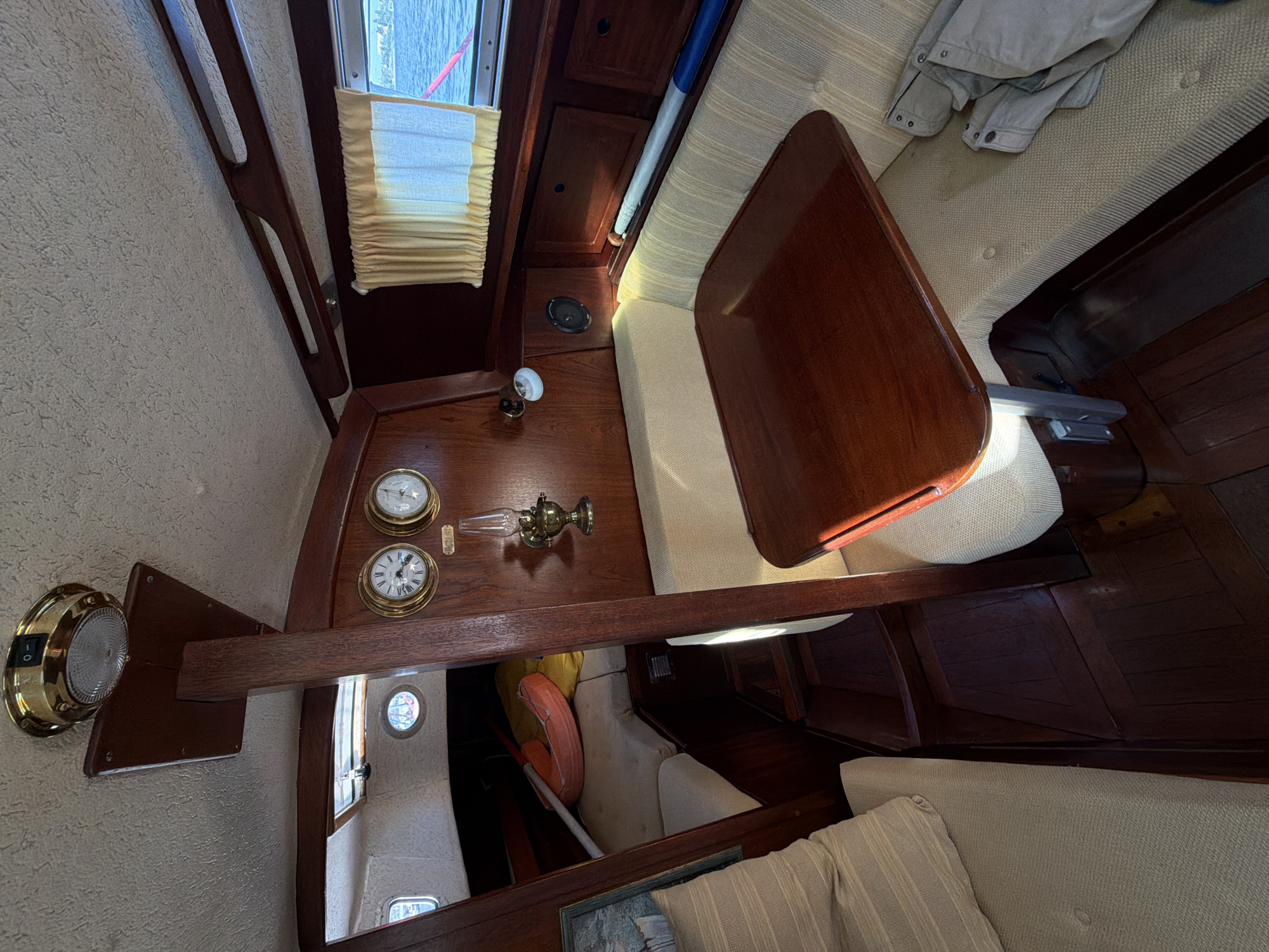 The image shows the interior of a sailboat's cabin. A wooden table with a polished surface is in the foreground, attached to a bench with upholstered seating. To the left, another section of seating is visible, with a pillow on it. The cabin walls and ceiling are made of wood paneling. A brass lamp is mounted on the wall, above two circular gauges, likely a clock and a barometer. A window with yellow curtains looks out onto a body of water and the sky. In the background, a life preserver and an oar are visible. There are no people present in the image. The image is undated and does not provide any information about its location.