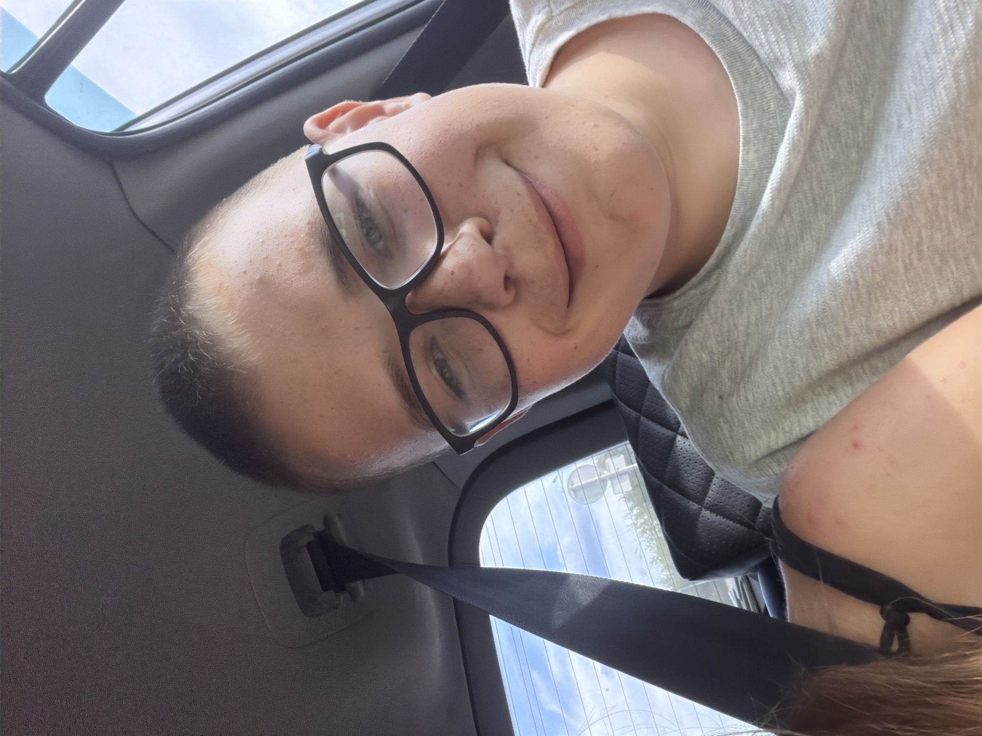 A young person with short hair and glasses is taking a selfie inside a vehicle. They are wearing a light gray t-shirt and have a slight smile. A seatbelt is visible on the left side of the frame, and the interior of the car, including the headrest of a seat, can be seen. Outside the car windows, a cloudy sky and trees are visible, suggesting a daytime setting. The specific location is Novoye Mavrino, Russia.