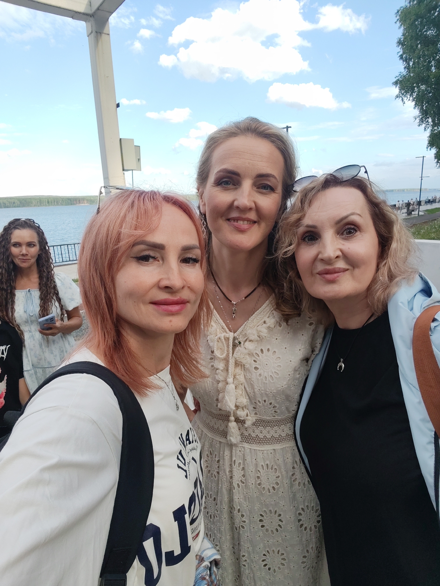 Three women pose for a selfie outdoors. The woman in the foreground on the left has light pink hair and is wearing a white t-shirt with dark lettering. She is wearing a backpack with black straps. To her right is a woman with blonde hair, wearing a white dress with intricate lace details and tassels. She has a necklace with a pendant. To the right of the blonde woman is another woman with blonde hair, wearing a black top and a light blue jacket. She also has a necklace with a crescent moon pendant. Her sunglasses are perched on her head. In the background, a fourth woman with long, dark curly hair is visible, wearing a light blue dress and holding a phone. She is standing near a railing. The scene appears to be a waterfront promenade, with a body of water, trees, and a clear blue sky with scattered clouds. The lighting suggests it is daytime, likely afternoon. People can be seen walking in the distance along the promenade. The overall mood seems casual and friendly. The visible text on the white t-shirt of the woman in the foreground appears to be a graphic design with some lettering, but it is difficult to read clearly.