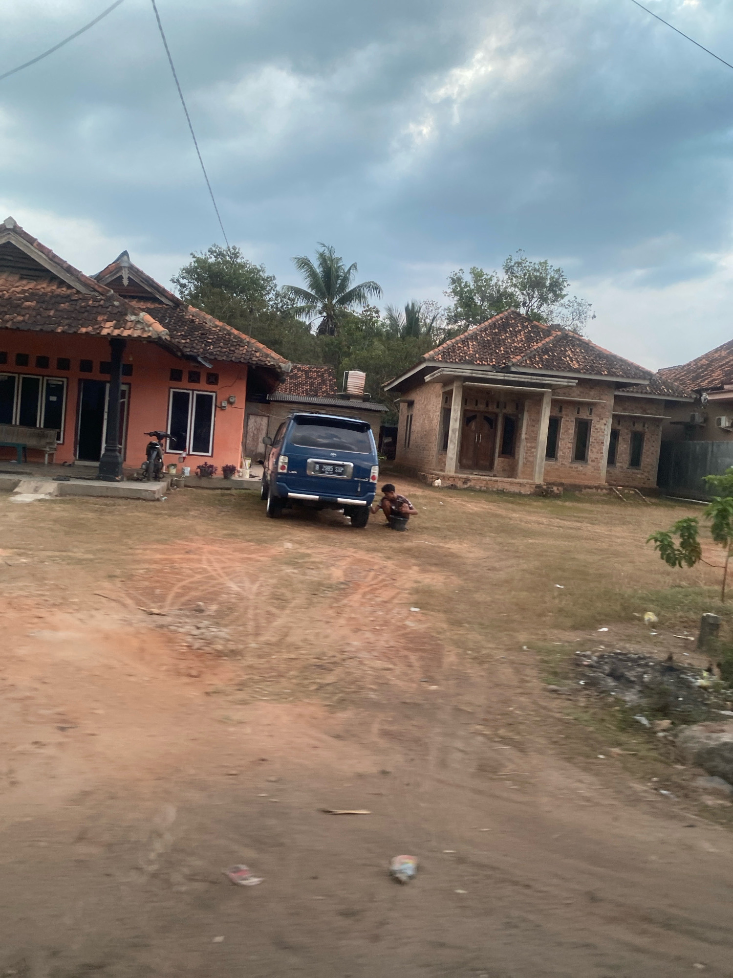 The image depicts a street scene in Toto Katon, Indonesia, under an overcast sky. In the foreground, a dirt road is visible, with scattered debris and vegetation. On the left side of the road, a single-story house with an orange facade and a terracotta tiled roof is situated. A motorcycle is parked on the concrete porch. To the right of this house, a blue minivan is parked. A person is crouched down beside the minivan, appearing to be engaged in an activity near the vehicle. Further down the road, another single-story house built with bricks and a terracotta tiled roof is visible. This house has a covered porch supported by pillars and multiple windows. In the background, trees and more houses are discernible.