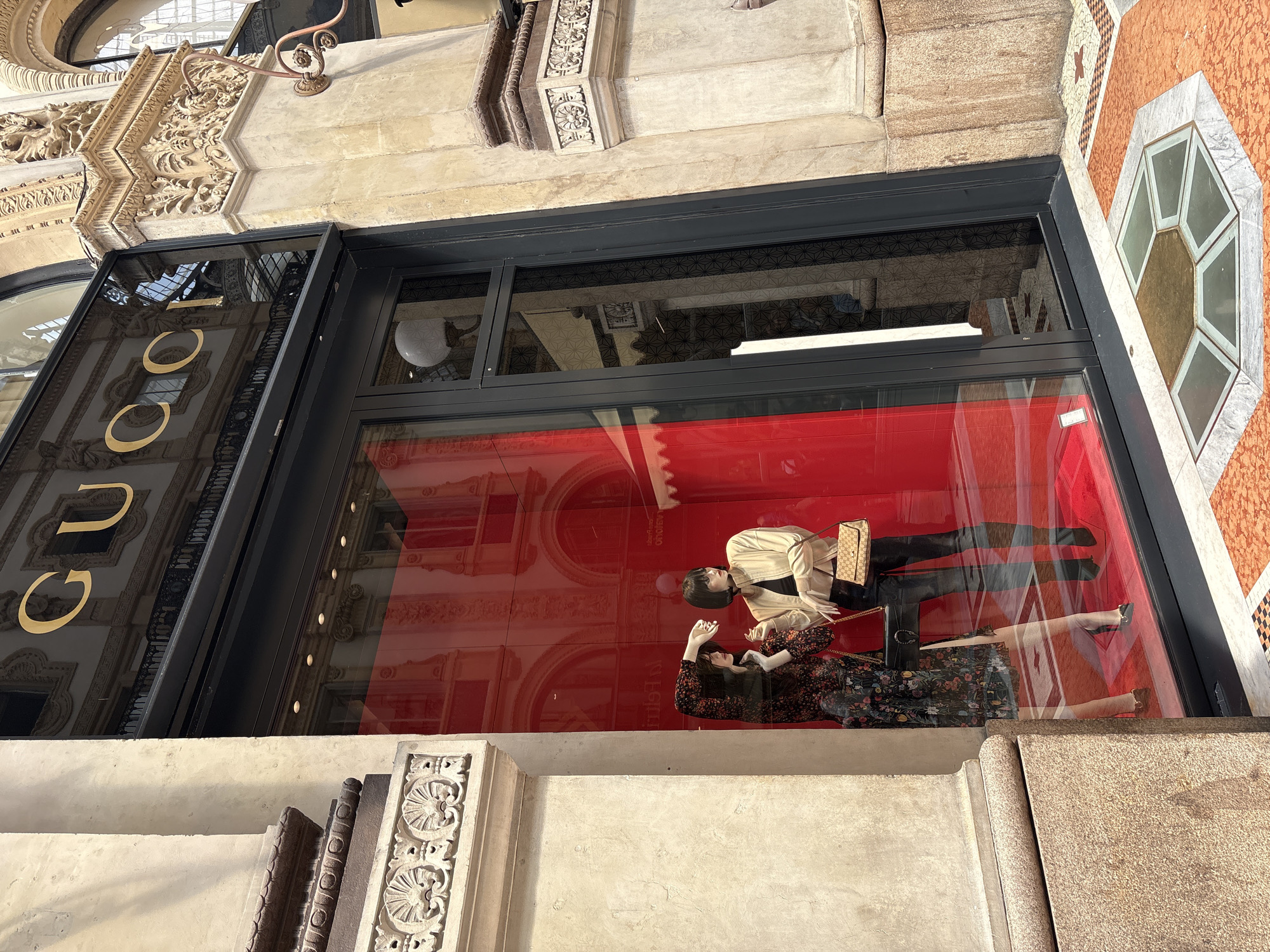 This is an image of a Gucci storefront in Milan, Italy. The building architecture is classic, with ornate carvings around the window frames. The storefront features a large window display with two mannequins. One is wearing a light-colored jacket and black pants, holding a purse. The other mannequin is wearing a floral dress and lying down. A red backdrop is present in the display. The Gucci logo is visible on the storefront. Reflections in the glass show the architecture of the building across the street. The scene is well-lit.