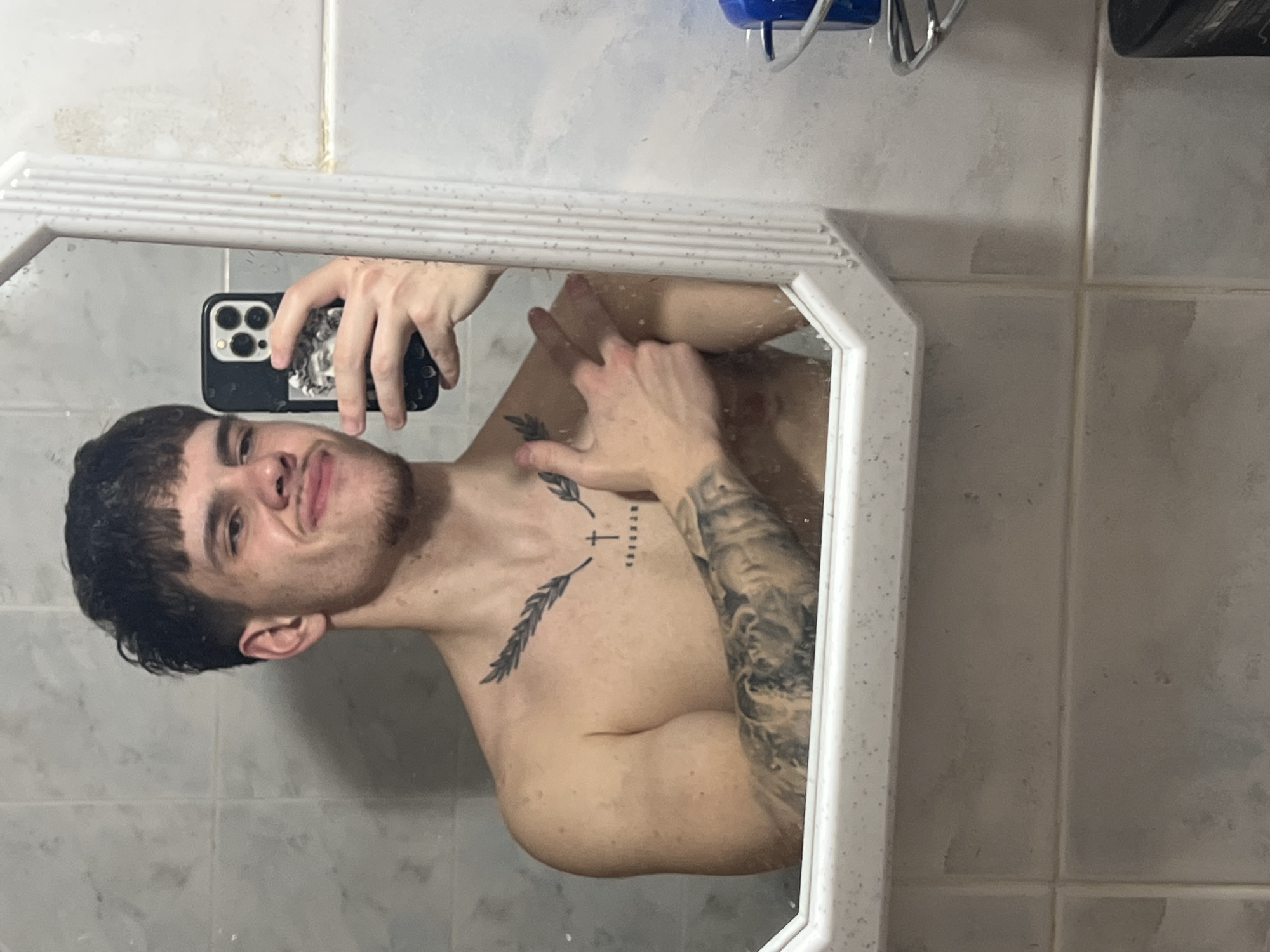 A shirtless man with short, dark hair is holding a smartphone in his left hand, taking a selfie in a white-framed bathroom mirror. The phone has a black case and multiple camera lenses visible. Tattoos are seen on the man's chest and left arm. The bathroom has white tiled walls. There is no clear indication of the location being in Sofia, Bulgaria or any specific city.