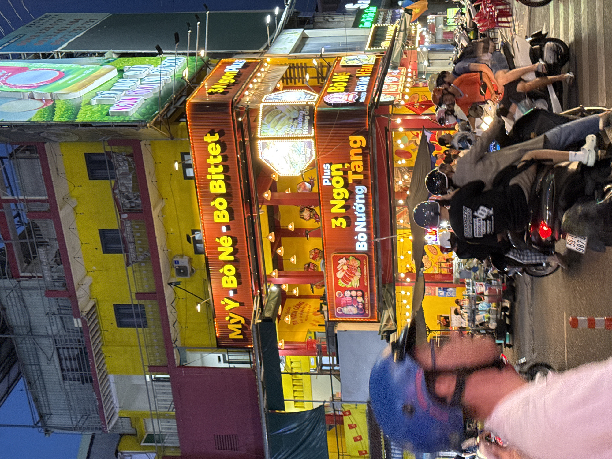 The image shows a street scene in Thủ Đức, Vietnam, at night. Brightly lit multi-story buildings with yellow and red facades are visible, adorned with large signs in Vietnamese advertising "Mỳ Ý - Bò Né - Bò Bittet" and "3 Ngonplus Bò Nướng Tảng." The street is filled with parked motorbikes and people, some of whom are seated on the motorbikes. In the foreground, a person wearing a blue helmet is partially visible. Streetlights and signage contribute to the well-lit atmosphere.