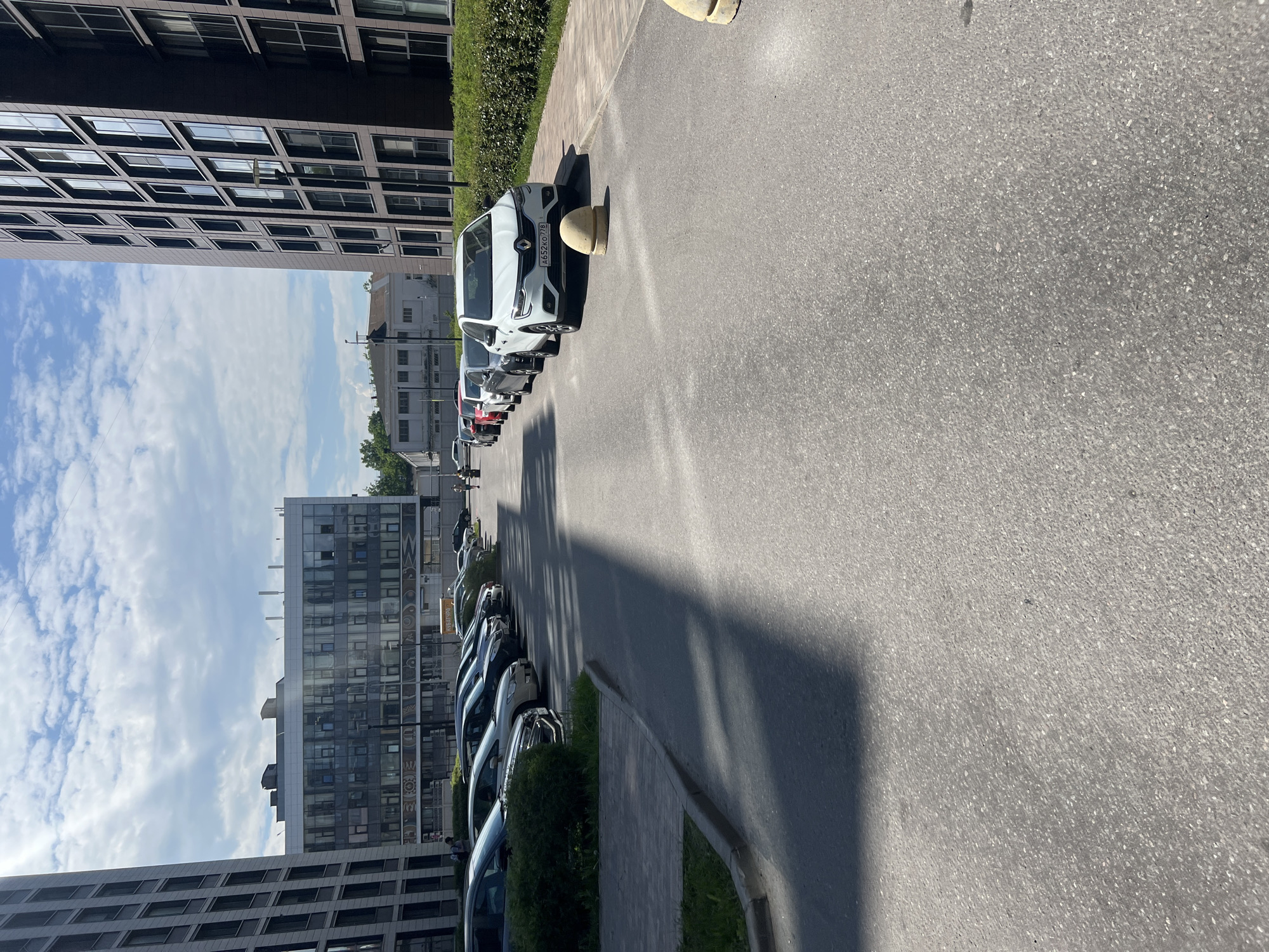 The image captures a bright, sunlit urban street scene in Saint Petersburg, Russia, likely in the midday or early afternoon due to the strong shadows cast across the asphalt.
The foreground is dominated by a wide, gray asphalt road stretching into the distance. To the left, a paved sidewalk borders a strip of low green bushes and a row of parked cars. The closest vehicle is a white van, possibly a Renault, identifiable by its Russian license plate "А652ХО 78", which indicates the Saint Petersburg region. This van is parked in front of a yellow concrete bollard. Behind it, several other cars are parked in a line along the sidewalk. Further down the street, another line of parked cars is visible on the right.
On the left side of the image, a large, modern building rises prominently, featuring a dark facade with a grid pattern of numerous rectangular windows reflecting the sky. The sky above is bright blue with scattered white, fluffy clouds. In the mid-ground, on the right side of the street, another multi-story building with a contemporary design, including large glass windows and some patterned decorative panels, stands. Parts of older, lighter-colored buildings are visible in the background under the clear sky.
A few indistinct figures of people can be seen in the distance near the buildings, but no specific activity or event is taking place; the scene is a static depiction of a typical city street with parked vehicles. The sunny weather and distinct shadows are notable details.