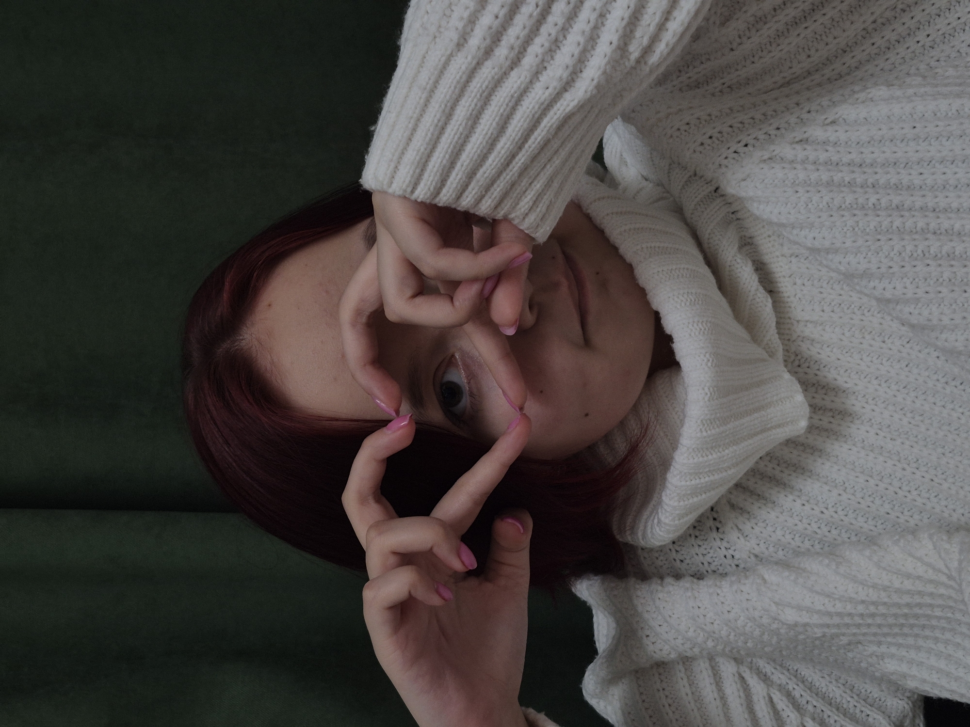 A person with auburn hair and pink nail polish is lying on a dark green surface, possibly a couch or bed. The person is wearing a chunky white knit sweater with a cowl neck. Their hands are positioned in front of their face, with fingers intertwined, partially obscuring their features. Only one eye is fully visible. The image is rotated counterclockwise, and the setting is indoors, but the location in the city of Chaikovsky, Russia, cannot be determined from the image alone.