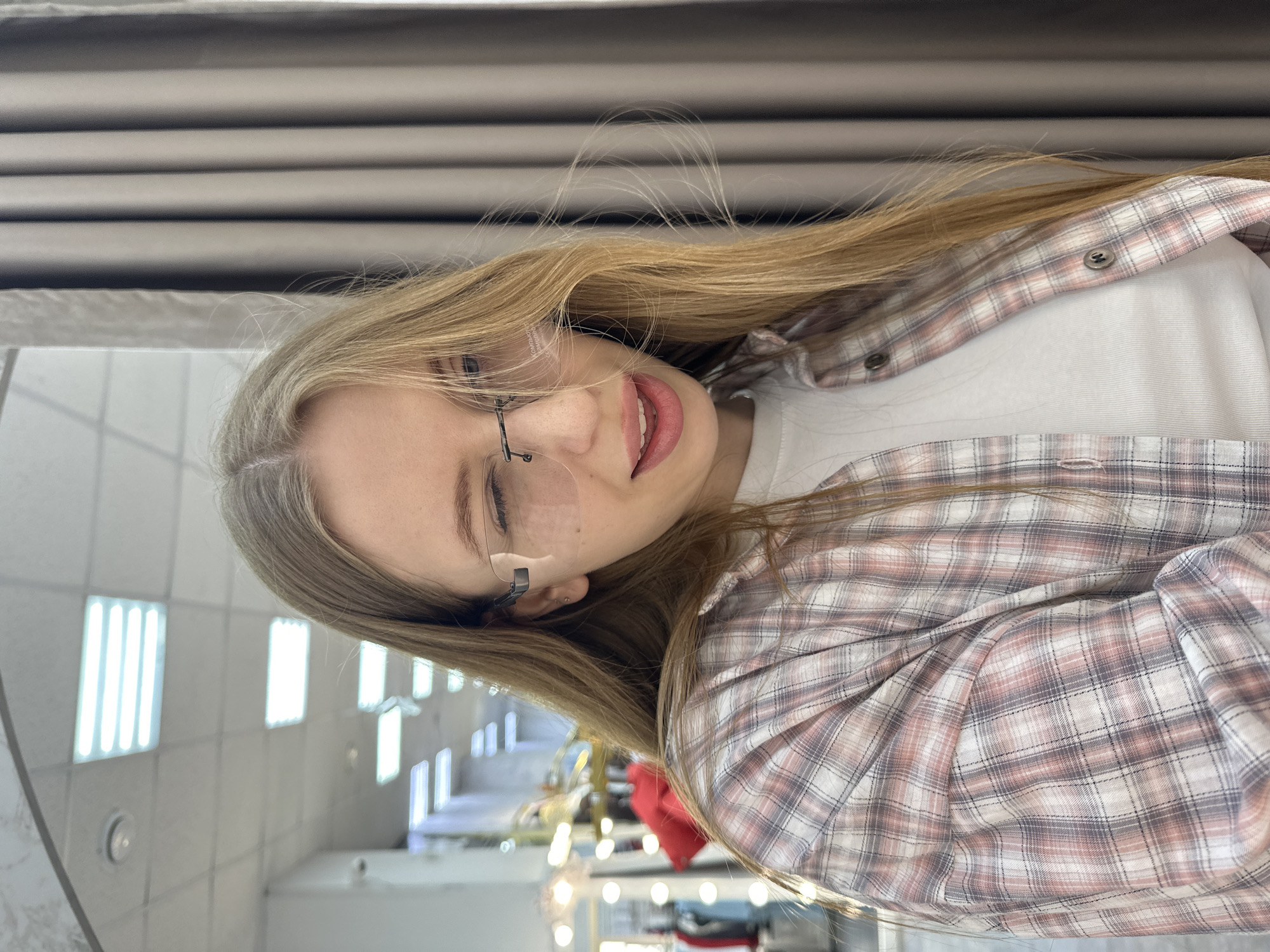 A young female, with long, light brown hair parted on her right, is depicted in a chest-up portrait within an indoor commercial setting in Chaikovsky, Russia. She is wearing rimless eyeglasses, a plaid shirt in shades of pink, white, and grey, unbuttoned over a white t-shirt. Her right eye is closed in a wink, and her mouth is slightly agape, revealing upper teeth. Two small stud piercings are visible on her left earlobe. The background on the viewer's left features a light-colored ceiling with multiple rectangular fluorescent light fixtures, a circular smoke detector, and a security camera on a white wall. Further left, blurred elements suggest clothing racks with garments, including a red item, and illuminated vanity mirrors. To the viewer's right, a beige curtain with vertical folds is visible.