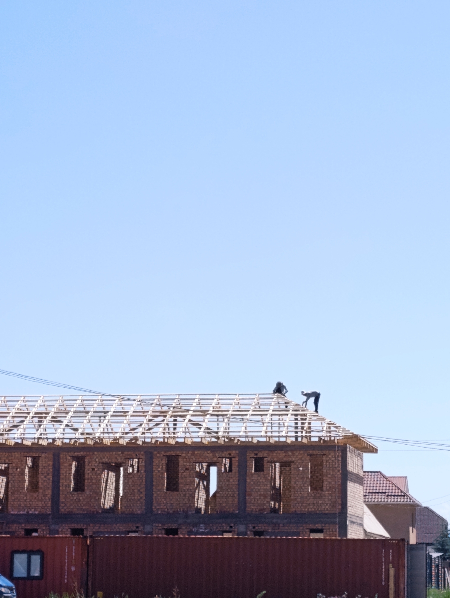 The image shows a construction site in Naryn City, Kyrgyzstan. Two construction workers are visible on the roof of a building under construction. The building appears to be made of brick and has an unfinished roof structure. In the foreground, there is a large, rusty metal container. The sky is clear and blue. Other buildings and a fence are visible in the background.