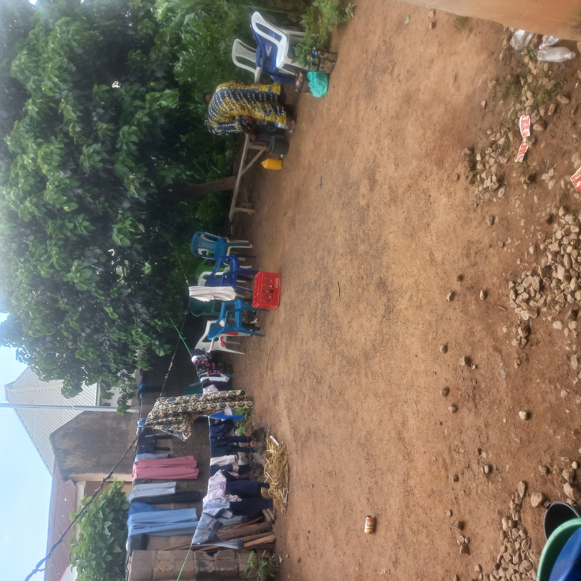 In Mangu Town, Nigeria, a rear yard scene unfolds with clothes drying on a clothesline strung between a brick wall and a tree. Various garments, including blue jeans, t-shirts, and dresses, hang, some with visible patterns. The ground is predominantly reddish-brown earth, scattered with rocks and sparse vegetation. In the foreground, a single beverage can lies on the dirt. Further back, plastic chairs of blue, white, and green are arranged near a red Coca-Cola crate. A woman, wearing a vibrantly patterned yellow and blue dress, is bent over, attending to something near a yellow container and a black bucket, with another person visible behind her. A wooden bench is positioned near a large tree. The environment suggests a typical residential outdoor space with signs of daily life.