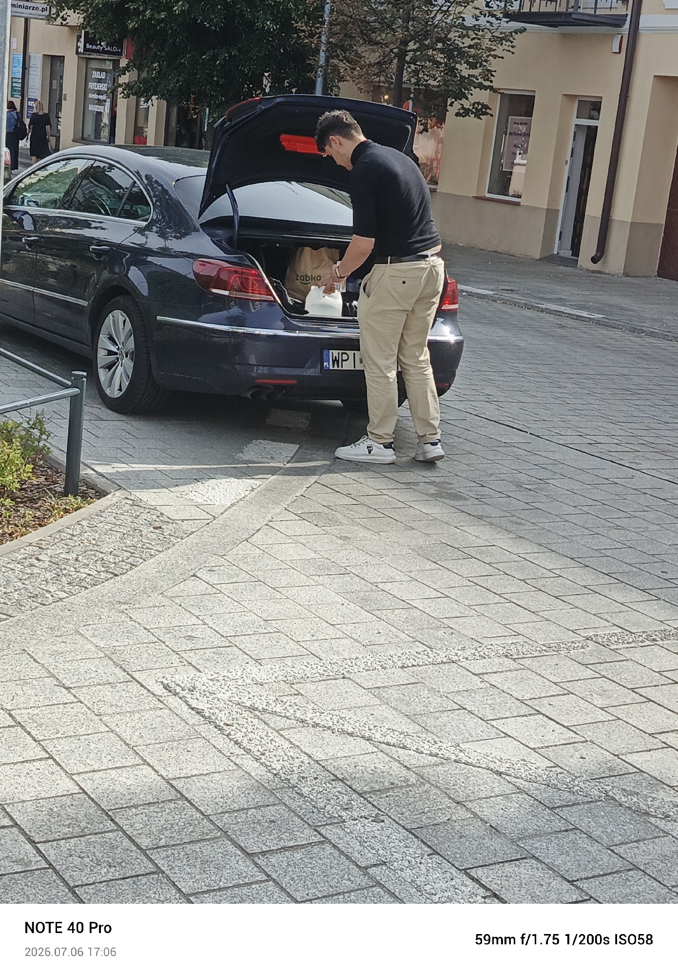 This media file depicts a man loading items into the trunk of a dark blue car, parked on a cobblestone street. The man is wearing a black t-shirt and light-colored pants, with white sneakers. He is holding a white grocery bag from "Żabka," a Polish convenience store chain, and appears to be placing it into the car. A partially visible license plate reads "WPI."
The scene takes place outdoors during the daytime, with bright sunlight casting shadows on the ground. In the background, there are buildings with storefronts, including one labeled "Beauty SALON" and another with "ZALAD FRYZJERSIO" and "MANICURE" visible. Trees line the street, suggesting an urban or suburban setting. The cobblestone pavement and the license plate's format (PL) indicate the location is likely in Poland. The timestamp at the bottom of the image reads "2026.07.06 17:06," which, given the current date, suggests this is a future date. The camera settings are also provided: "59mm f/1.75 1/200s ISO58." The overall impression is a candid moment of someone completing a task, possibly grocery shopping, on a sunny day in a Polish town.