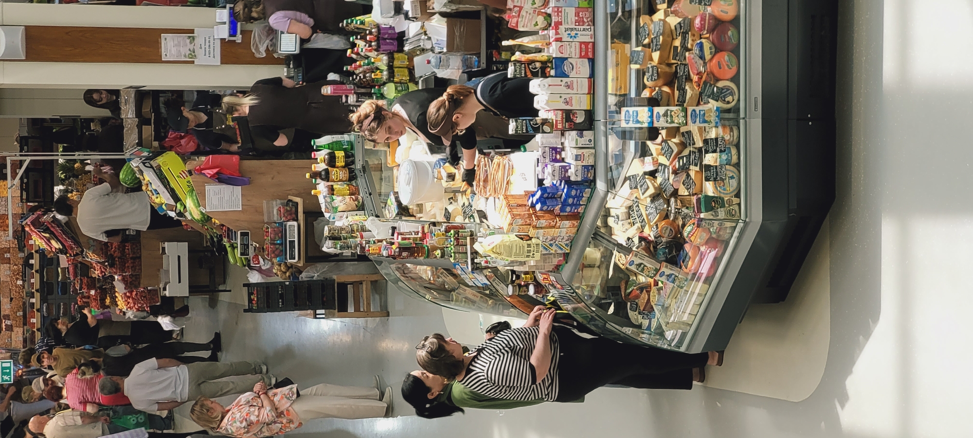 The image depicts an indoor multi-level market hall in Unknown, Unknown.
In the foreground, a curved, refrigerated display case filled with a variety of cheeses in blocks, wedges, and wheels, many with visible price tags, occupies the lower right. On top of this counter, numerous packaged dairy products, including milk and yogurt cartons, are stacked alongside assorted bottles of sauces, oils, and other condiments. A white plastic bucket rests on the counter's left side. Two female customers stand at this counter on the left; one wears a dark green top, and the other wears a black and white striped t-shirt and black pants. Behind the counter, two female staff members are present. One staff member, with dark hair in a bun and wearing a dark top with a brown apron and black gloves, is leaning over the display, appearing to serve or arrange items. Another staff member, wearing glasses and a dark top, stands to her right, observing the counter.
In the mid-ground and background, a bustling market floor is visible. To the left, a woman in a floral shirt and light trousers stands, facing right. On the upper level, a fruit and vegetable section features large quantities of produce, including strawberries in clear containers, watermelons, and bananas, displayed on wooden stands. Staff members in dark aprons are visible behind these stalls, assisting customers. A man in a white t-shirt stands at a fruit stall on the right. Further into the background, additional market stalls and patrons are seen, with shelves displaying various goods. A green exit sign with a white arrow is mounted on a wall in the upper left background. On the far right, behind a white pillar, two more female staff members are visible behind a counter that includes a cash register. The floor is a light, speckled grey.
