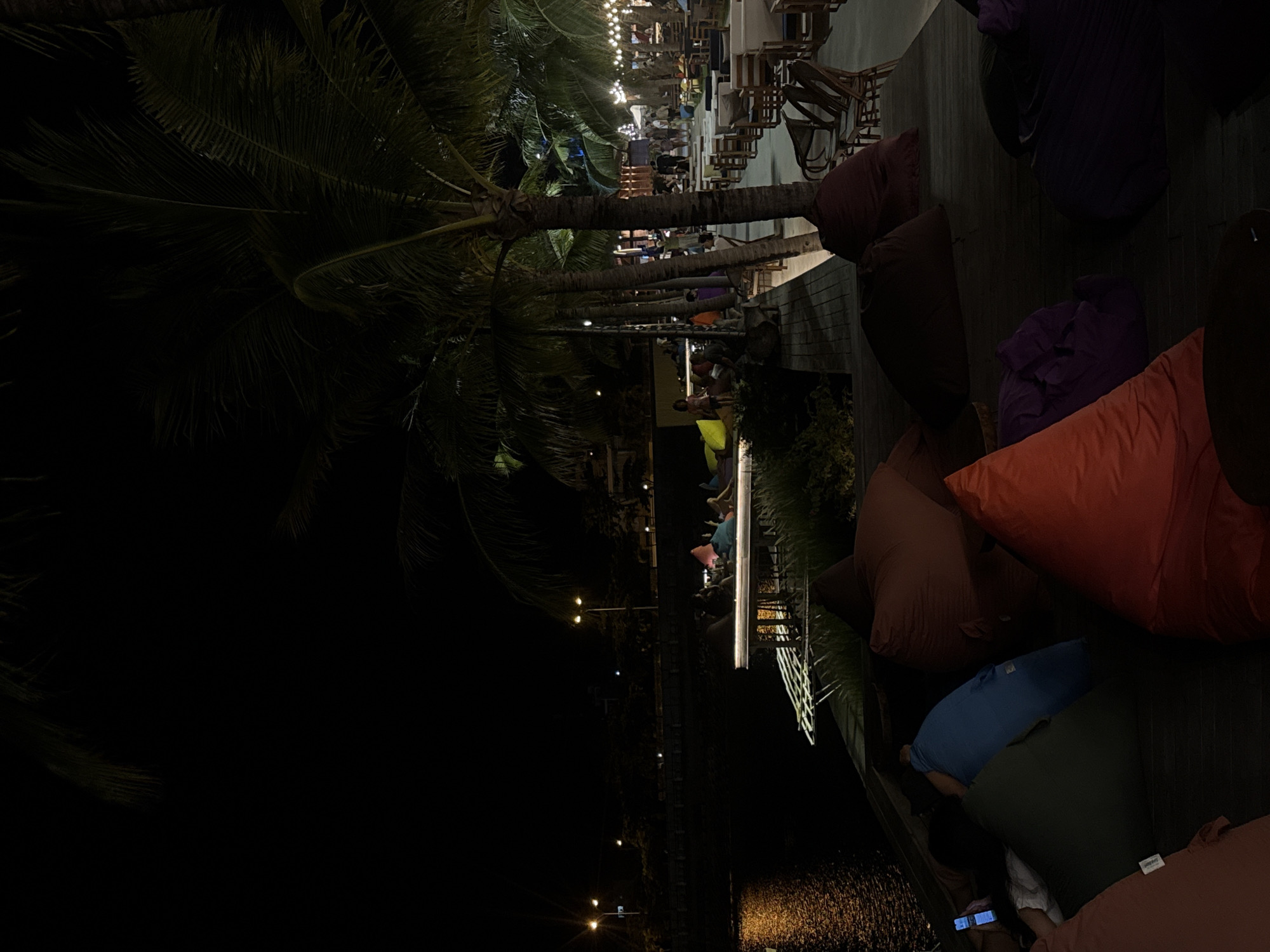 The image depicts an outdoor seating area at night, likely a restaurant or cafe, located on a wooden deck adjacent to a body of water. Palm trees are visible in the background, suggesting a tropical or subtropical location. The scene is illuminated by string lights and streetlights, casting a warm glow on the environment. Several people are seated on beanbag chairs and at tables, appearing to be engaged in conversation or enjoying the ambiance. The water surface reflects the lights, creating a shimmering effect. The overall atmosphere is relaxed and inviting, indicative of a leisure setting.