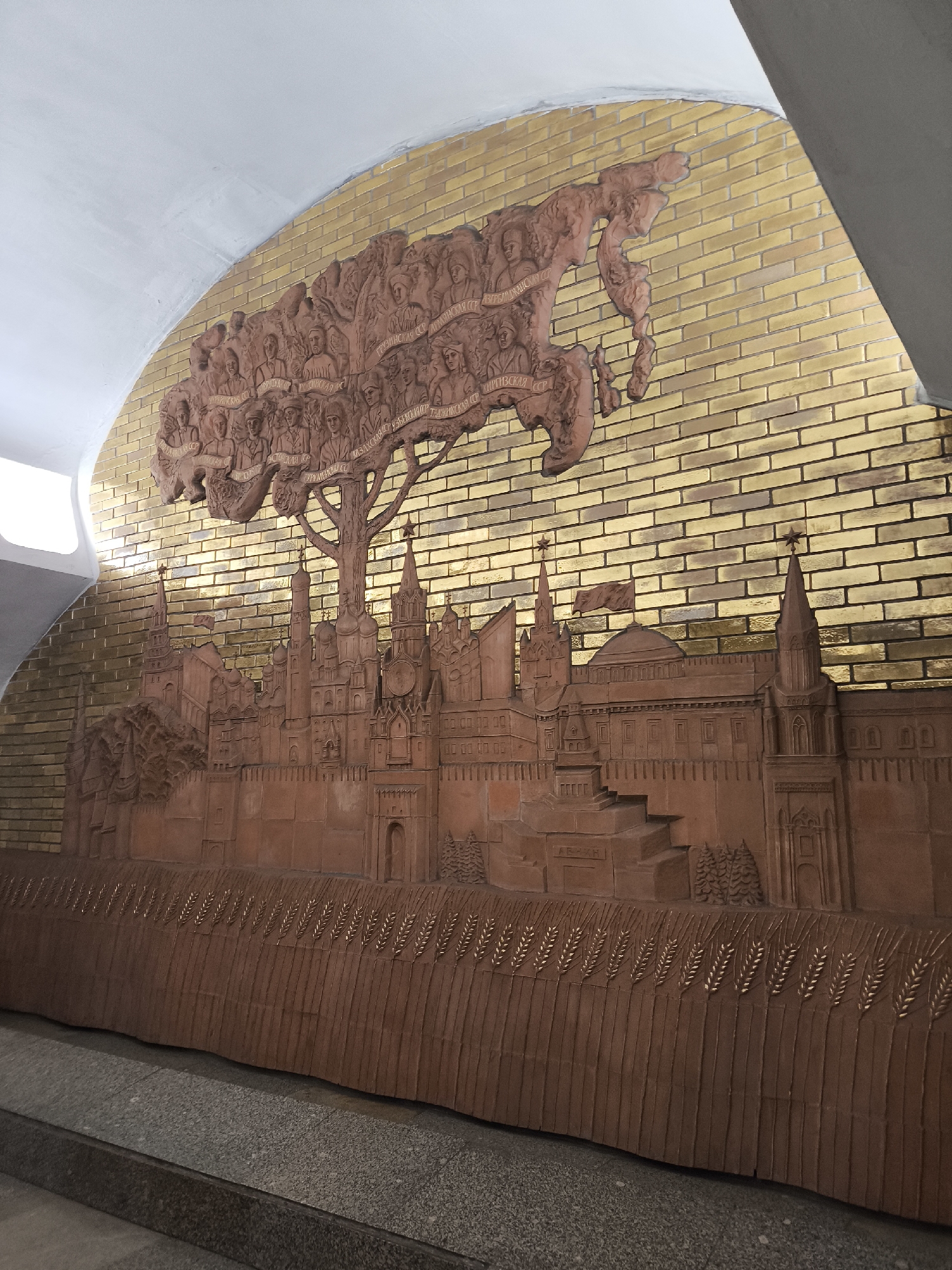 A low-angle, close-up shot presents a large, terracotta-colored relief mounted on a gold-toned, brick-patterned wall under an arched ceiling. The relief depicts a skyline of the Kremlin in Moscow, Russia, recognizable by its iconic towers and walls. Above the Kremlin, a tree-like formation is adorned with portrait busts and place names in Cyrillic. Below the Kremlin, stylized wheat stalks are horizontally arranged along the base of the relief. The bottom part of the image is the gray floor. The city is Unknown, Unknown.