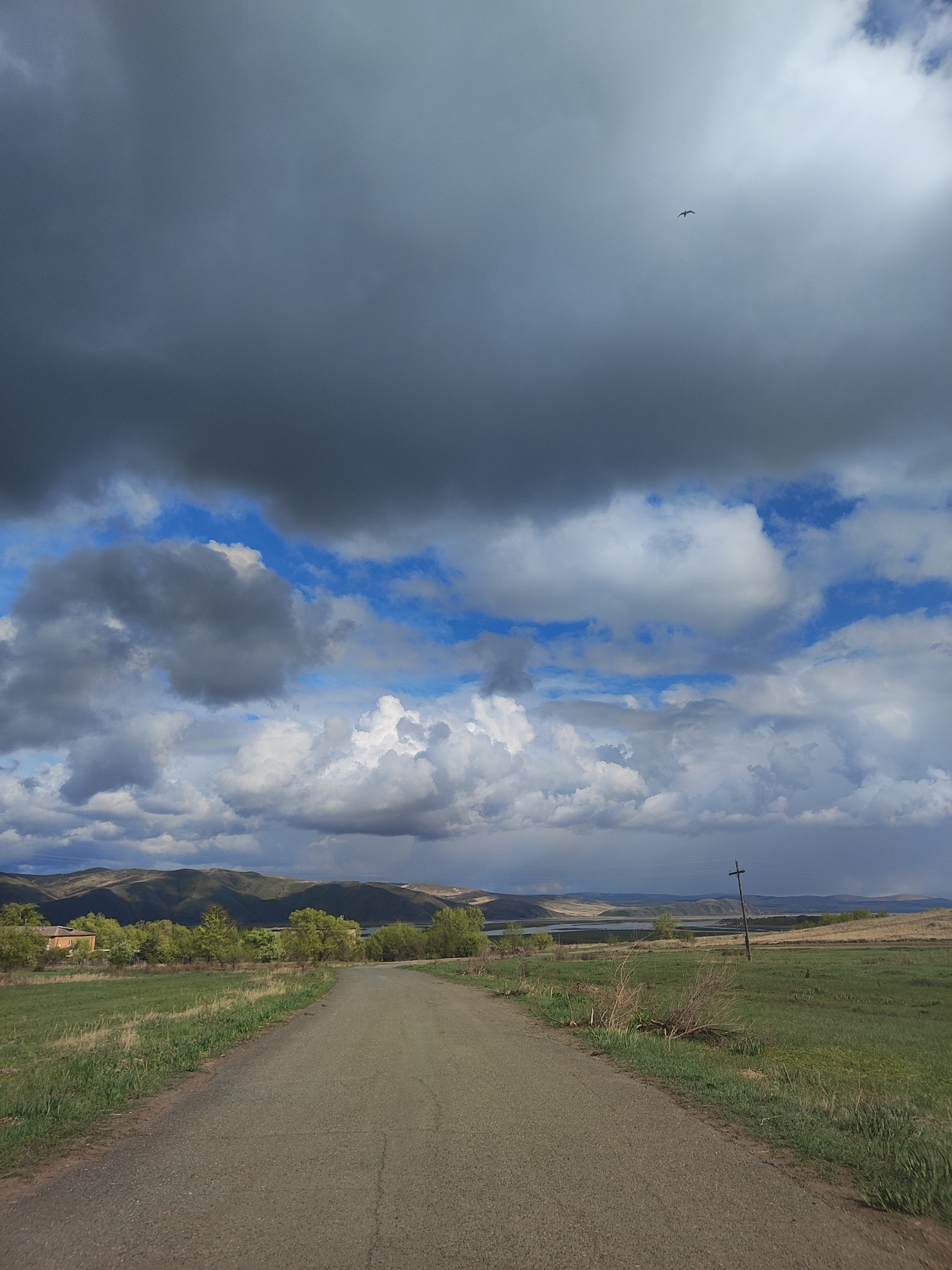 A rural road stretches forward in the Unknown landscape. The road, appearing as if made of dirt or gravel, leads towards distant hills under a dramatic sky. Dark, ominous clouds dominate the upper portion of the sky, contrasting with patches of blue sky and white, fluffy clouds. A single bird is visible in the distance against the cloudy sky. Along the road's sides, green fields of grass are visible, with some vegetation encroaching on the road's edges. A lone wooden power pole stands to the right of the road. In the distance, a few buildings and trees can be discerned, with hills and a body of water visible behind them. The overall environment suggests a remote or countryside location.