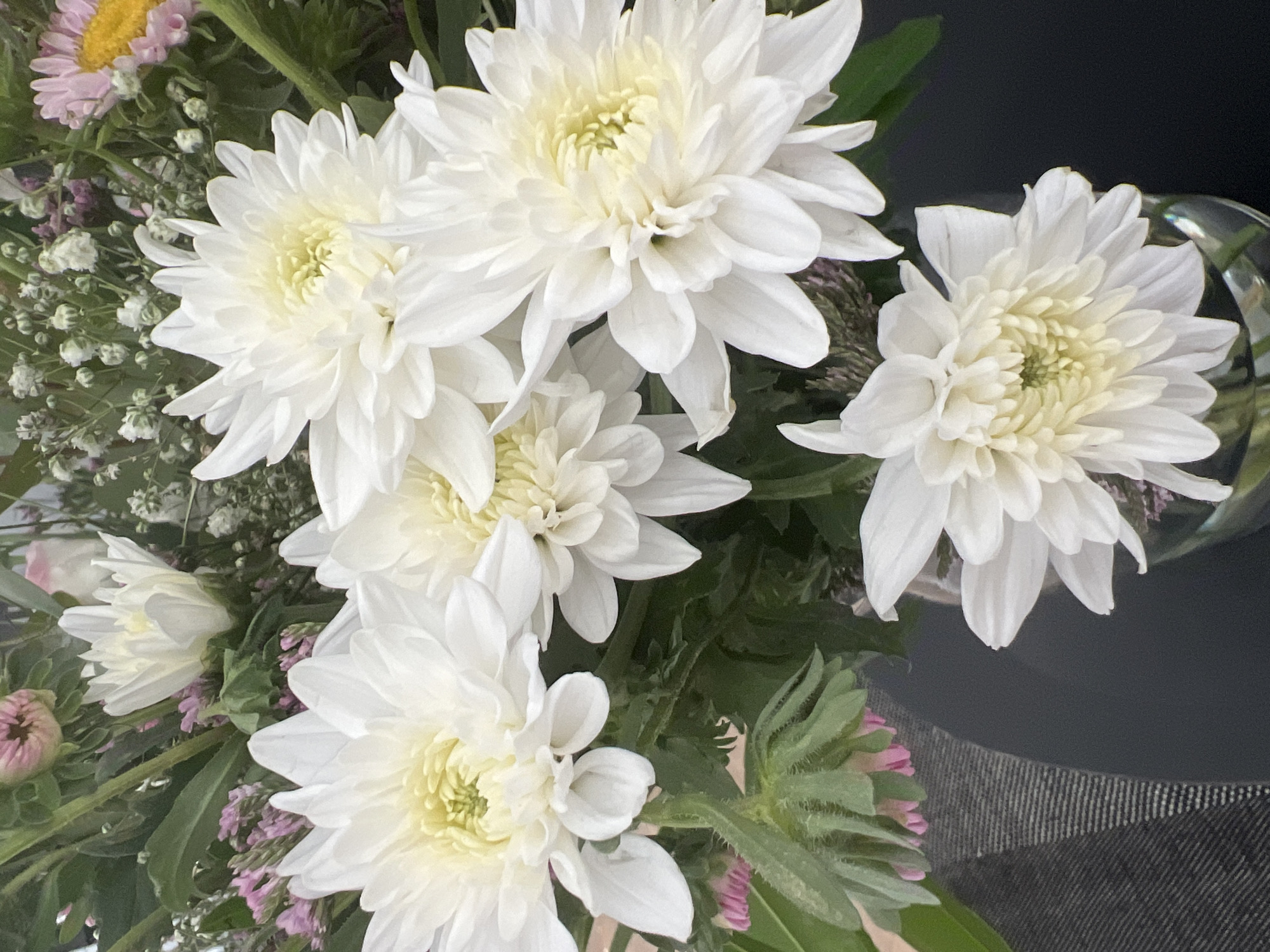 A close-up, overhead shot captures a bouquet of white chrysanthemums and various small filler flowers, possibly gypsophila and statice, arranged in a glass vase. The chrysanthemums are the dominant feature, with their layered white petals and pale yellow centers. Some are in full bloom, while others appear partially open. In the background, hints of pink and yellow daisy-like flowers are visible. The bouquet is placed on a dark, textured surface, likely a table, and a portion of a gray, striped fabric is visible in the lower left corner. The lighting is soft and natural, illuminating the delicate details of the petals and foliage. The scene is located in the city of Hof HaCarmel Regional Council, Israel. There are no people visible in the image.