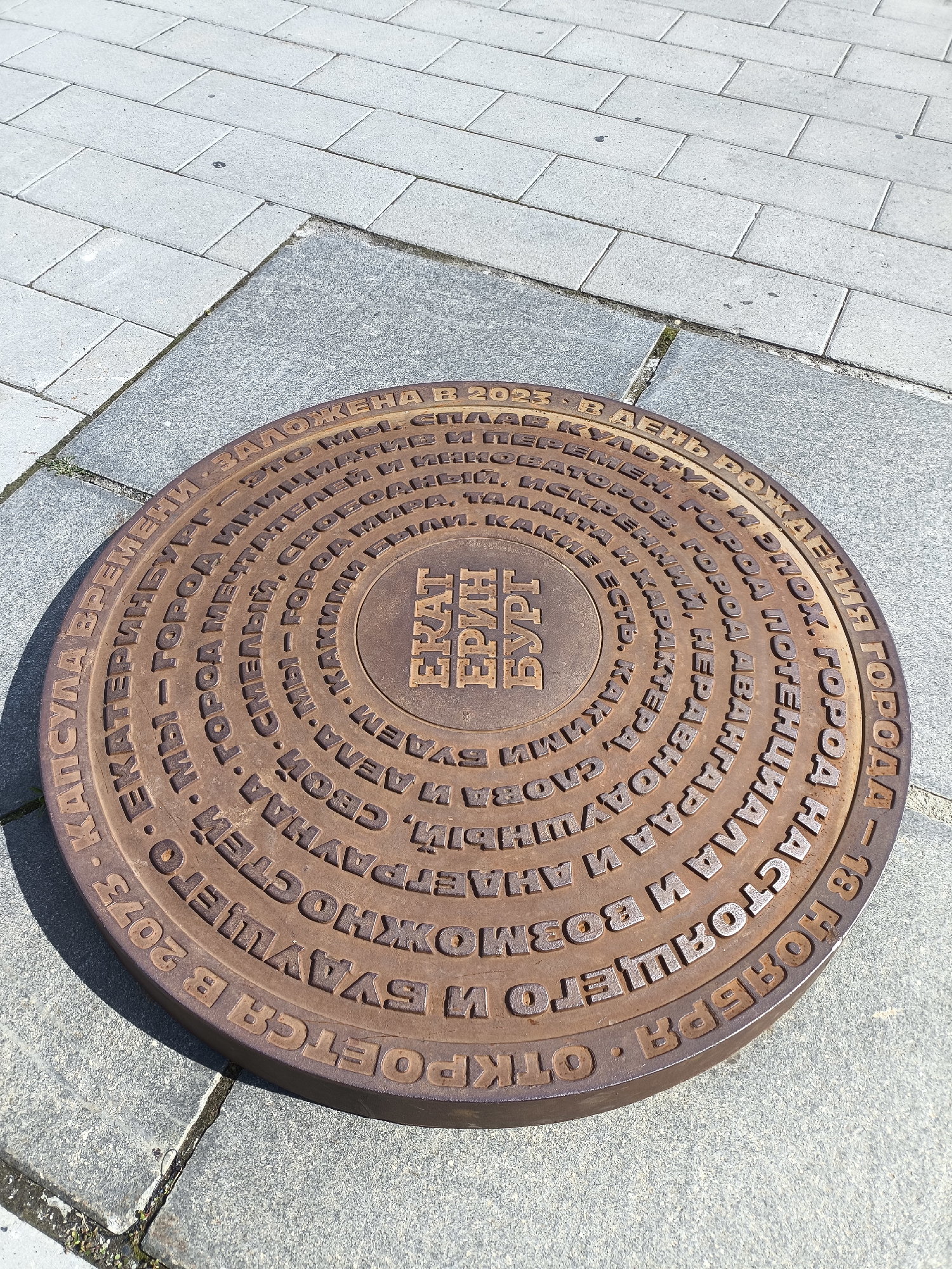The image shows a circular metal manhole cover with Russian text inscribed on it, set into a paved sidewalk. The manhole cover is made of a reddish-brown metal and has a concentric ring design with text in raised letters. In the center of the manhole cover is a square emblem with the text "ЕКАТЕРИНБУРГ" in stylized letters. The surrounding text on the manhole cover is a poem or inscription about the city of Yekaterinburg. The sidewalk is made of light gray rectangular paving stones and darker gray granite-like stones. The scene is outdoors, under bright sunlight, as indicated by the shadows cast by the manhole cover and the overall bright illumination. The manhole cover is the primary subject, with its inscription being the most notable feature. There are no people or animals visible in the image. The environment is urban, with paved ground. The inscription on the manhole cover reads, in part: "КАПСУЛА ВРЕМЕНИ - ЕКАТЕРИНБУРГ - ГОРОД МЕНЯЮЩИЙ СВОЙ ОБЛИК. КАКИМИ МЫ БУДЕМ В 2073 ГОДУ? ОТКРОЕТСЯ БУДУЩЕГО И БУДУЩЕГО И ВОЗМОЖНОСТЕЙ СВОИМ СЛОВОМ И ДЕЛОМ. НАСТОЯЩЕГО НАСТОЯЩЕГО. 18 НОЯБРЯ - В ДЕНЬ КУЛЬТУРЫ РОЖДЕННАЯ ГОРОДА - ЕКАТЕРИНБУРГ". This translates to: "TIME CAPSULE - YEKATERINBURG - A CITY CHANGING ITS APPEARANCE. WHAT WILL WE BE LIKE IN 2073? THE FUTURE AND THE FUTURE AND POSSIBILITIES WILL BE OPENED BY ITS WORDS AND DEEDS. OF THE PRESENT OF THE PRESENT. NOVEMBER 18 - ON THE DAY OF CULTURE BORN OF THE CITY - YEKATERINBURG".